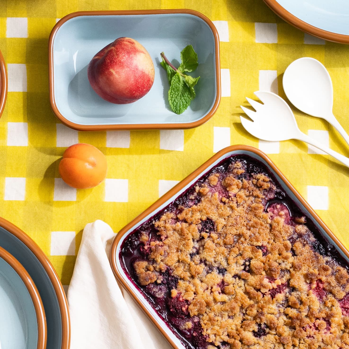 An overhead view of enamel baking dishes filled with food, enamel serving set, and a napkin on a bright yellow crosshatch tablecloth