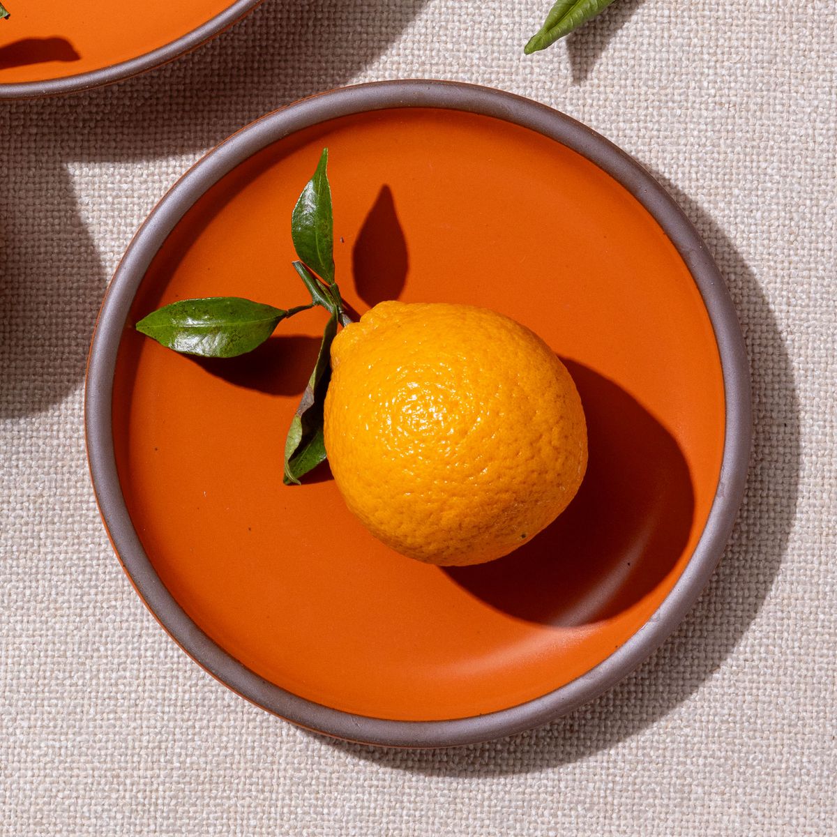An orange sits on a dessert sized ceramic plate in a bold orange color featuring iron speckles and an unglazed rim
