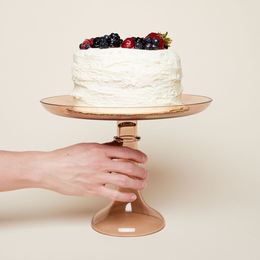 A hand touches an amber glass cake stand with a tall white cake on top