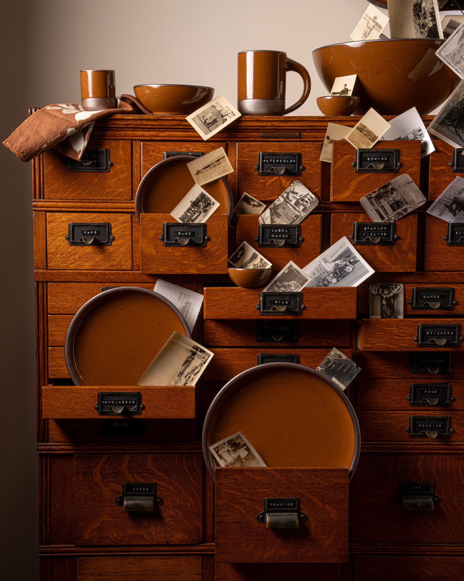 Glossy caramel brown ceramic mugs, bowls, and plates displayed in a wooden drawer cabinet with scattered vintage photos.