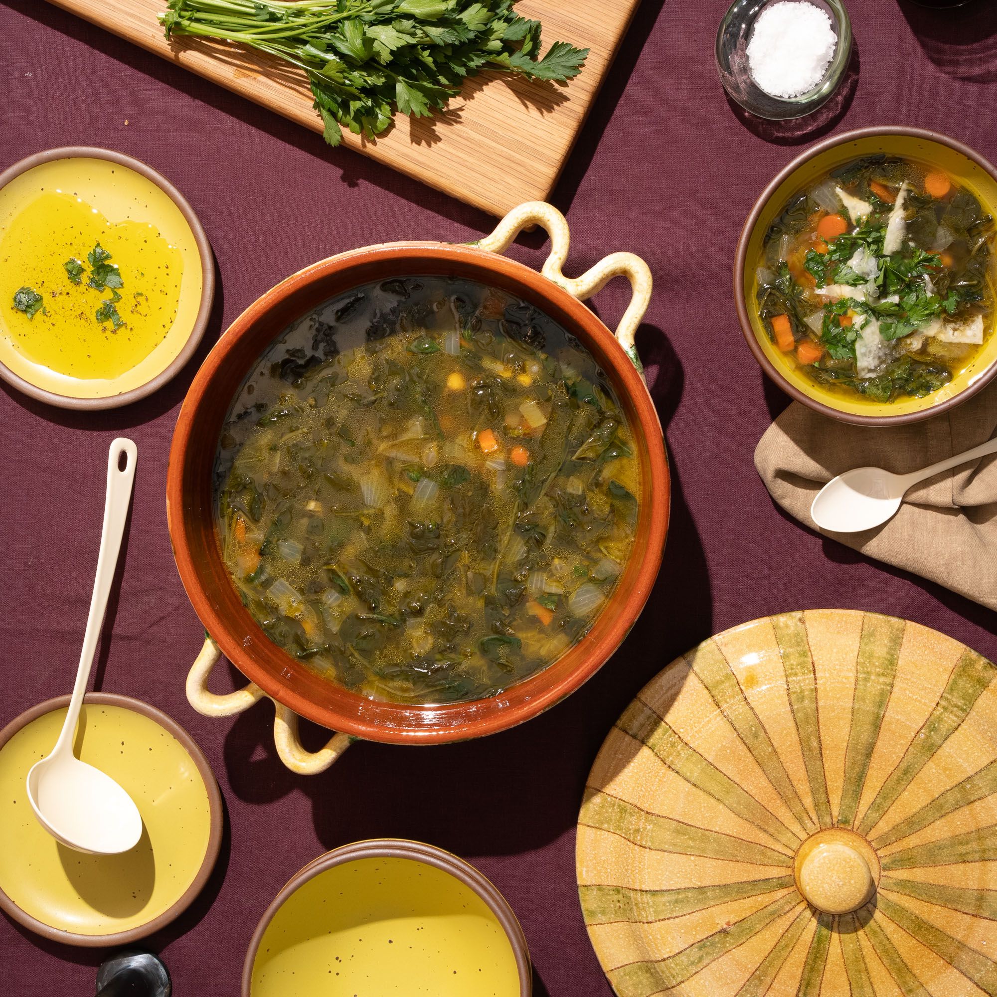 A bird's eye view of a terracotta casserole dish with soup inside and the lid off to the side, with yellow ceramic soup bowls and plates, a ladle, and more.