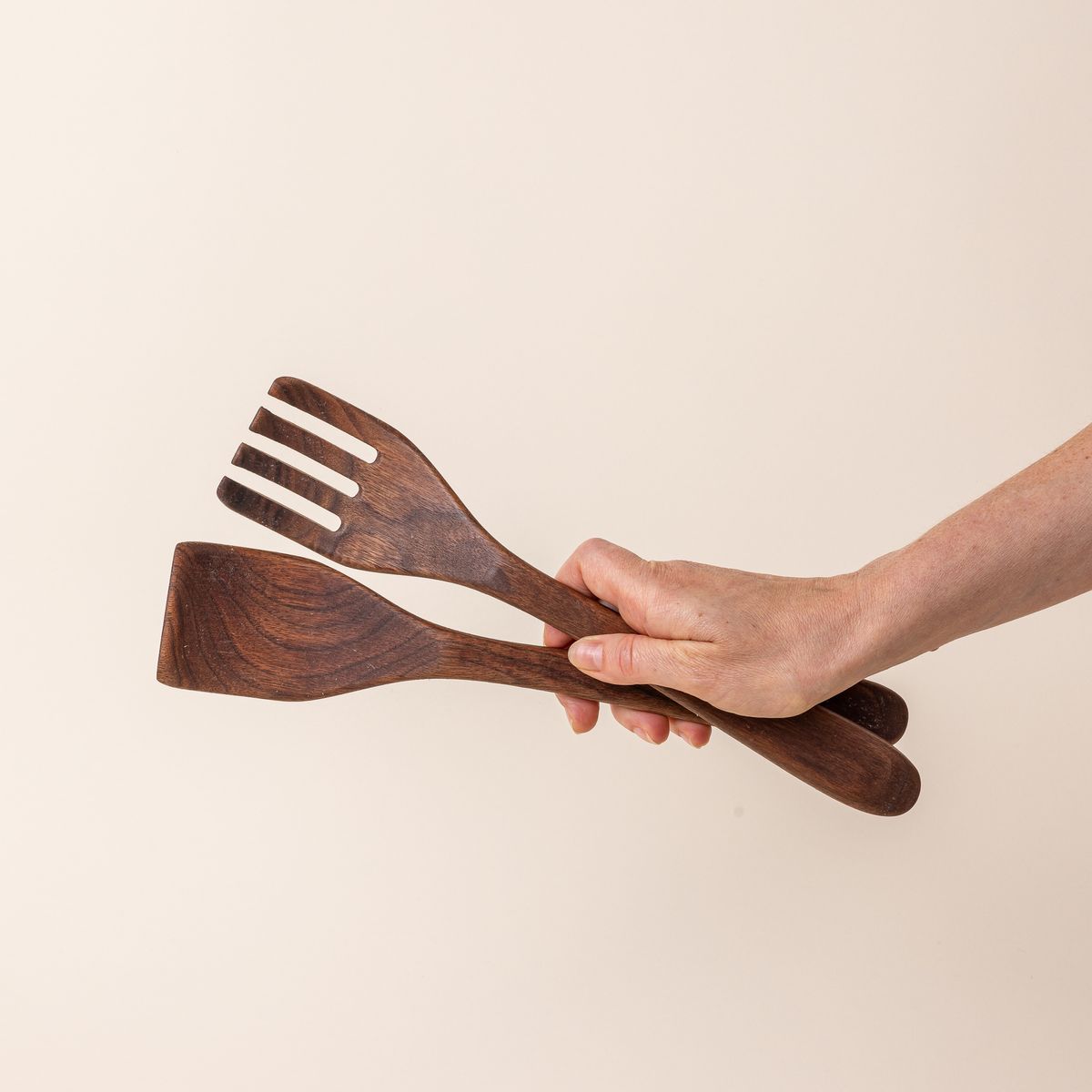 Hand holding a pair of long walnut wood serving utensils. 