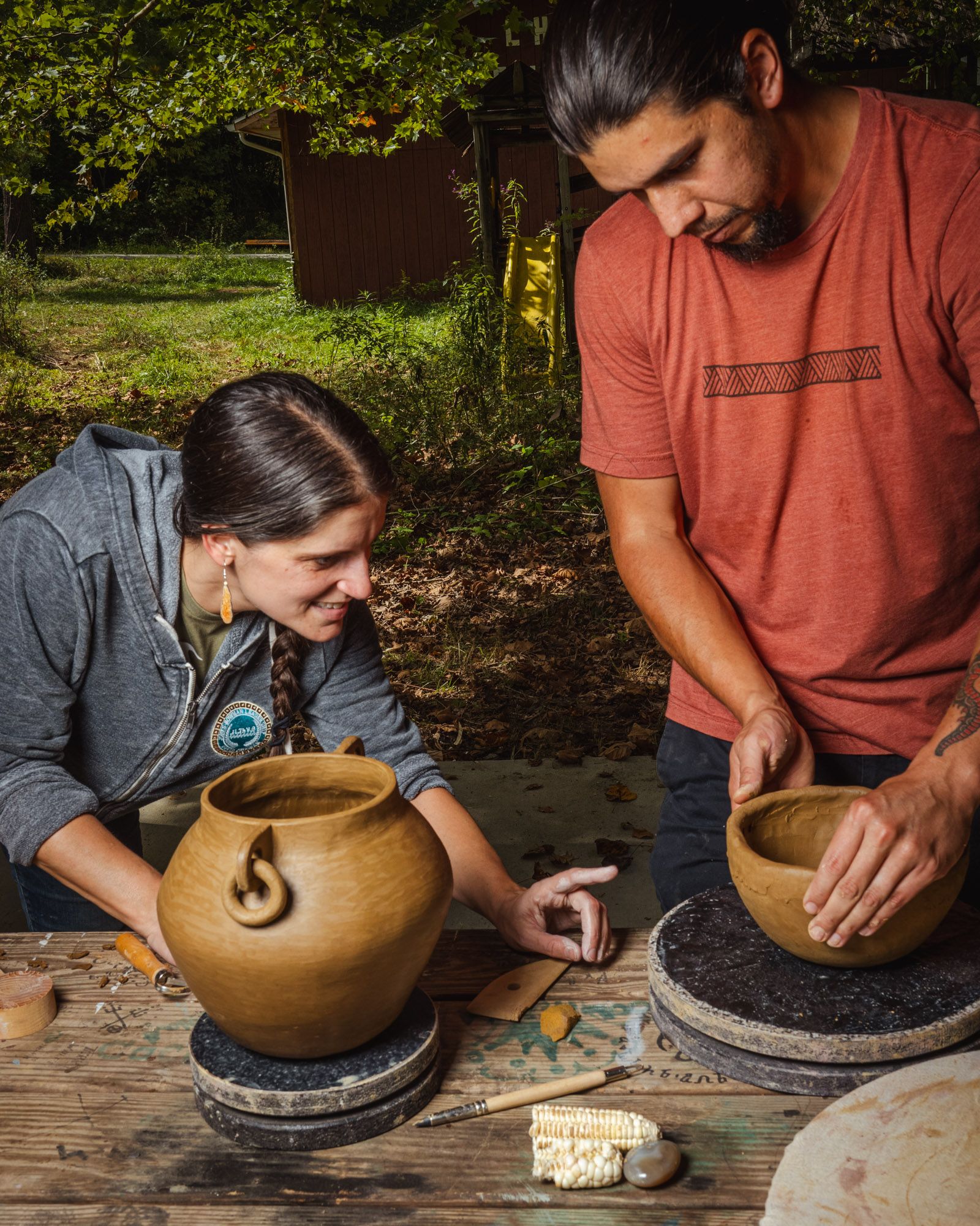 Two people working on creating and forming clay pots at a table with 1 person bending and pointing at a clay bowl while another person forms the shape