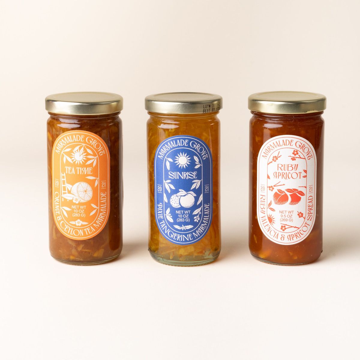 Three jars of Marmalade Grove spreads: Orange & Ceylon Tea Marmalade (orange label), Sunrise Pixie Tangerine Marmalade (blue label), and Ruby Apricot Spread (red label).