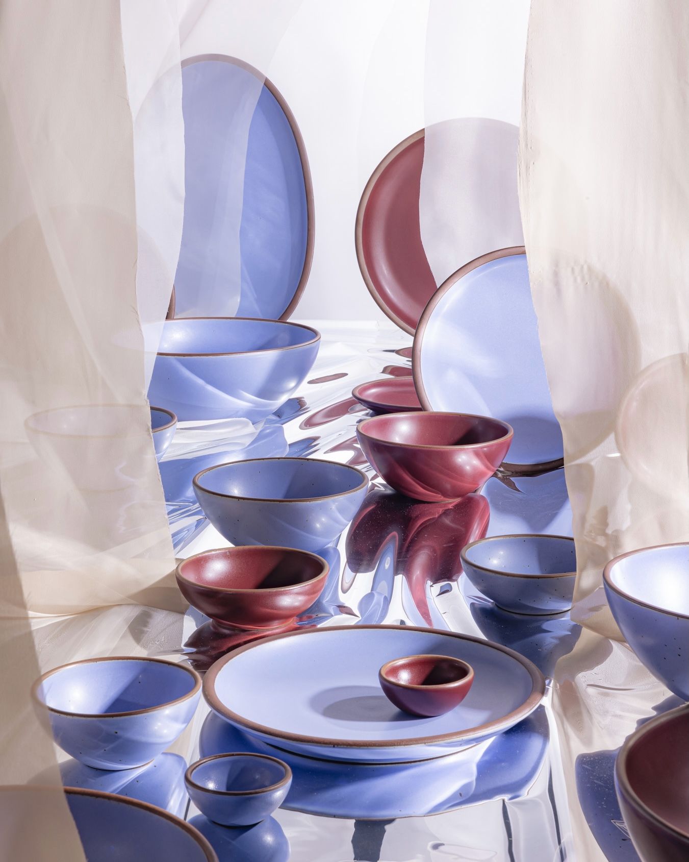 Ceramic plates and bowls in periwinkle and plum colors are artfully arranged against fabric and a reflective material