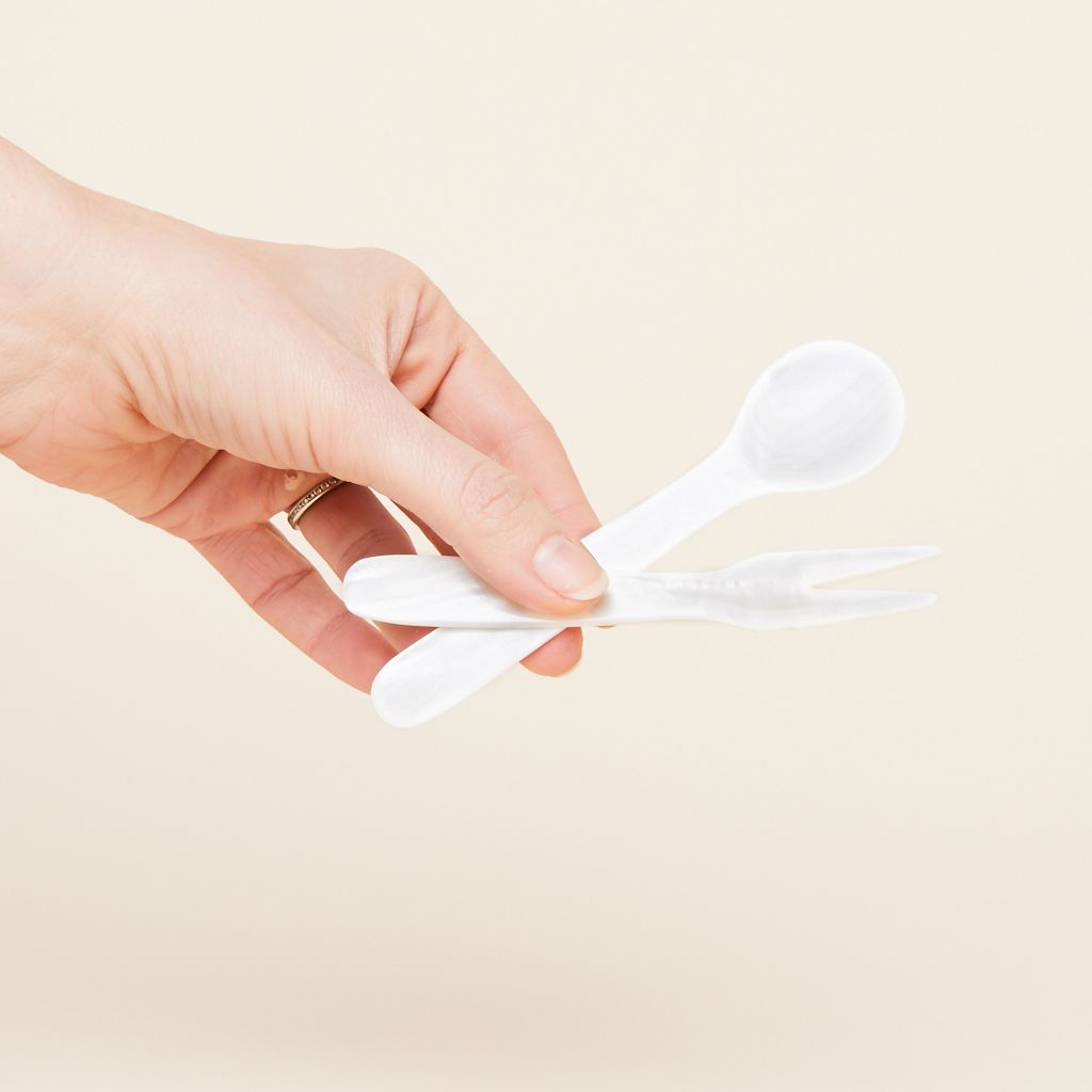A hand holds a white spoon and a white fork, both of which are made of shell