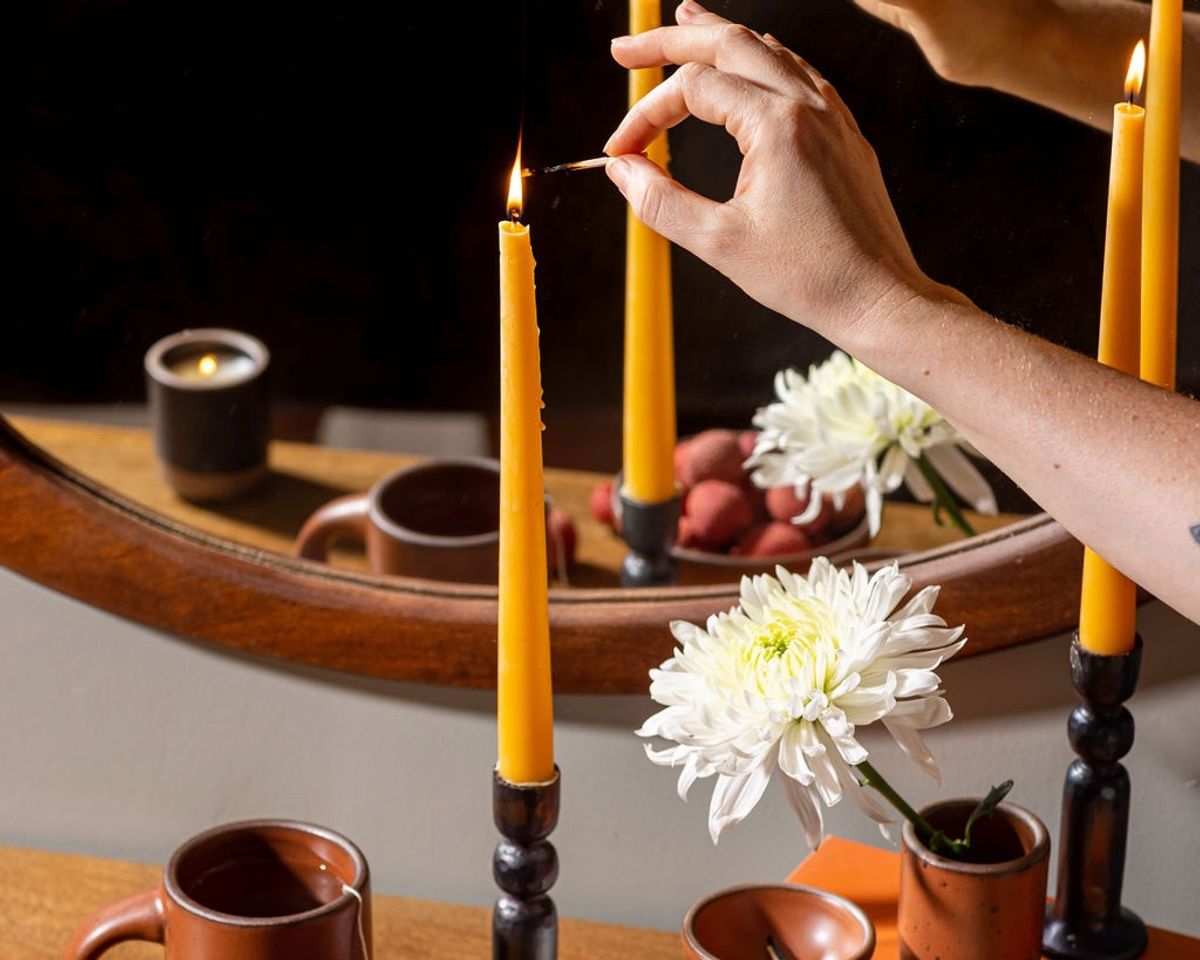 A hand lights a taper candle in an iron candleholder. Nearby is a small cup with a large flower, an Amaro East Fork Mug, and a circular mirror in the background.
