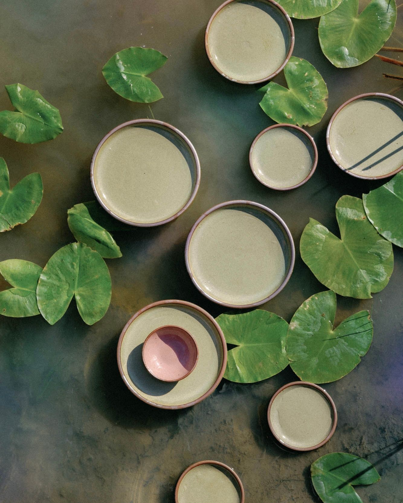 An overhead view of reflective dappled grey-green and dusty mauve pink plates and bowl floating in a pond with lily pads.
