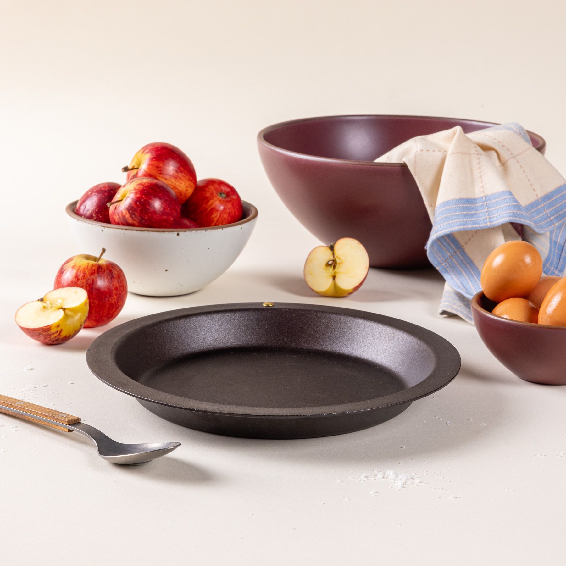 A simple iron pie dish with a wide rim and shallow body, surrounded by large ceramic bowls with apples and eggs.