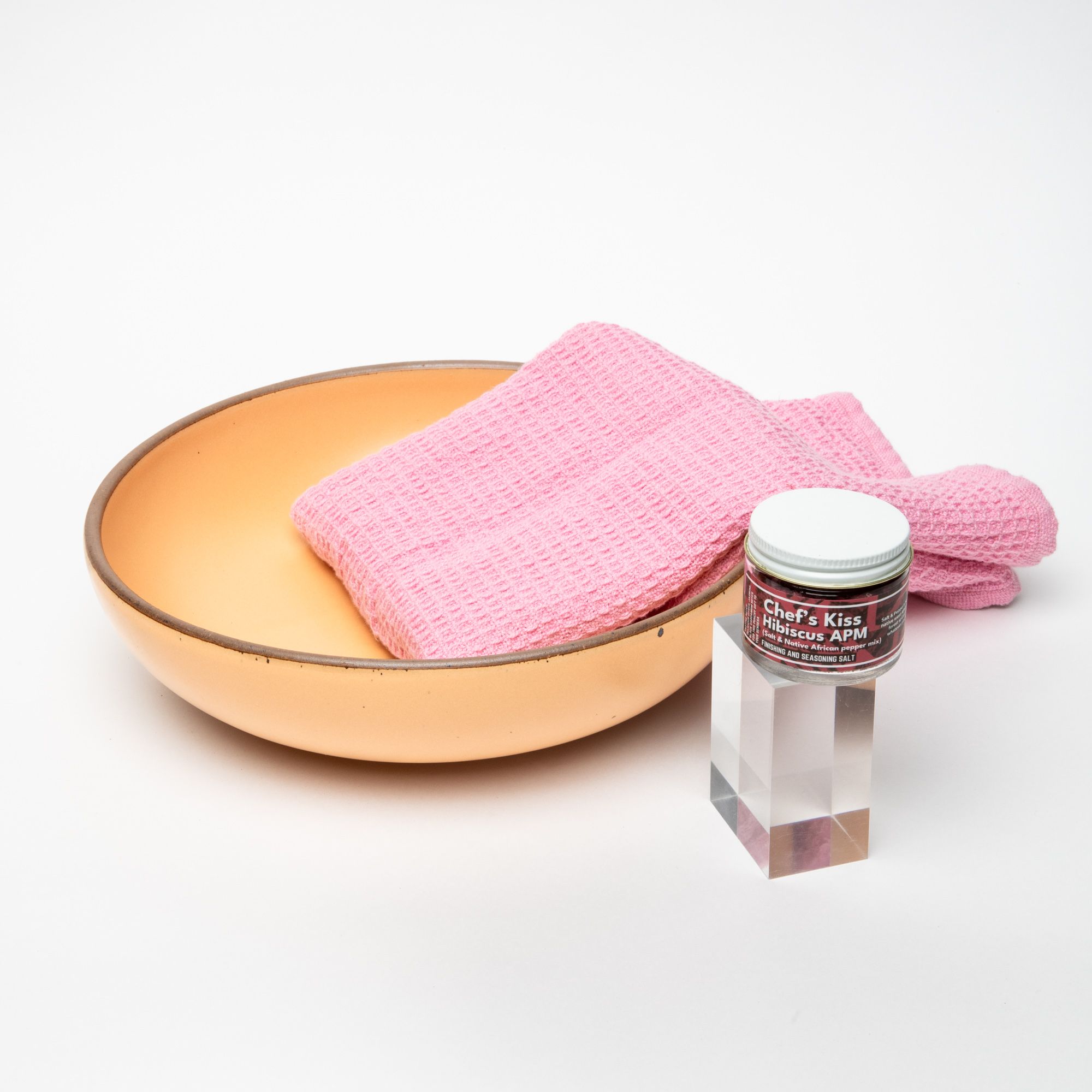 A large serving bowl in a peachy yellow color with a warm pink waffle towel and a small jar with a white top and a label that reads "Chef's Kiss Hibiscus APM"