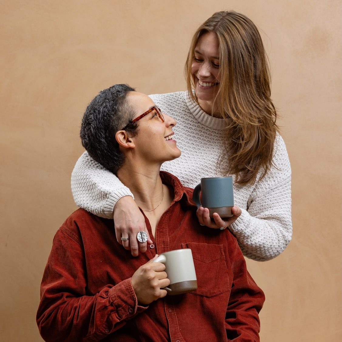 A couple is smiling and close together holding ceramic mugs in white and blue-grey colors.