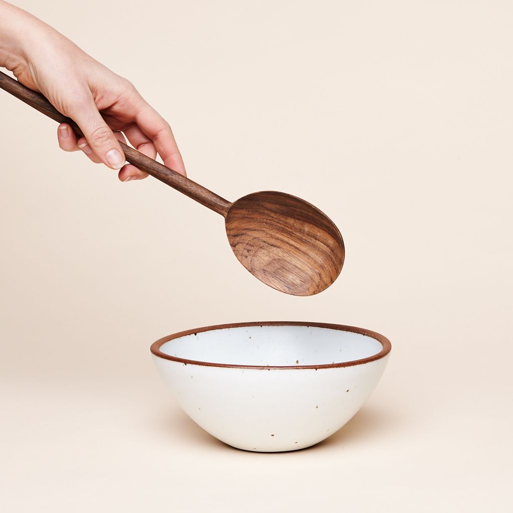 A hand holds the handle of a long, dark brown wooden spoon with a pattern in the grain over a white bowl with a brown rim.