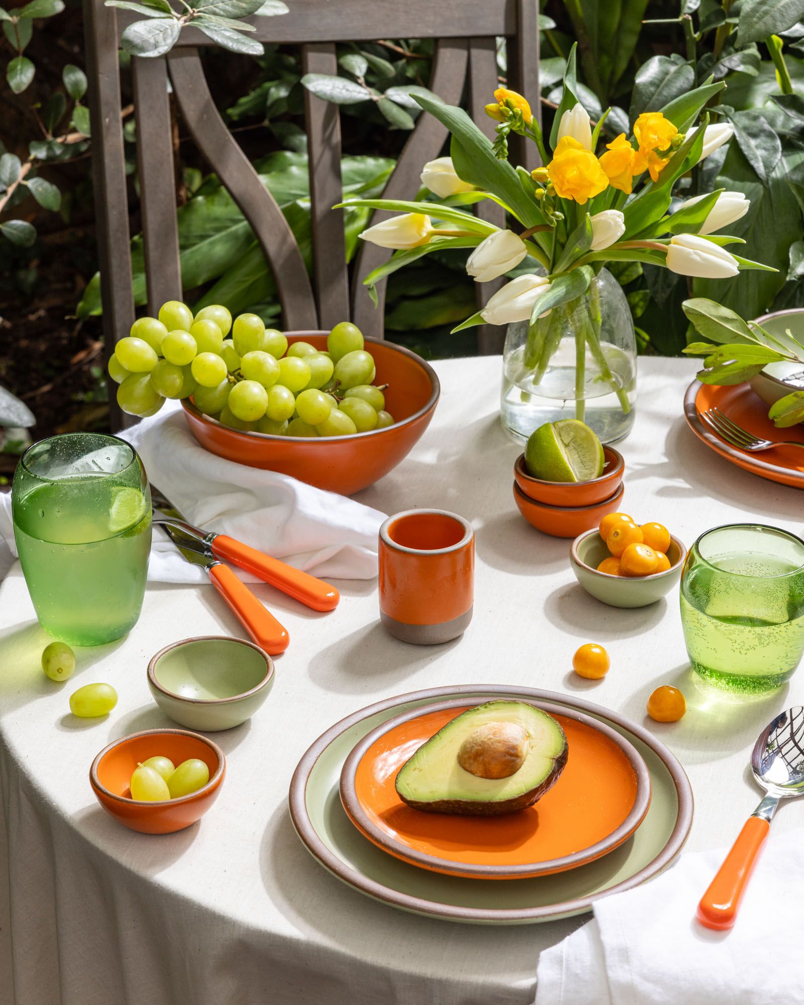 An outside table filled with ceramic plates and bowls in a bold orange and calm sage colors, breakfast foods, orange flatware, and green glassware.
