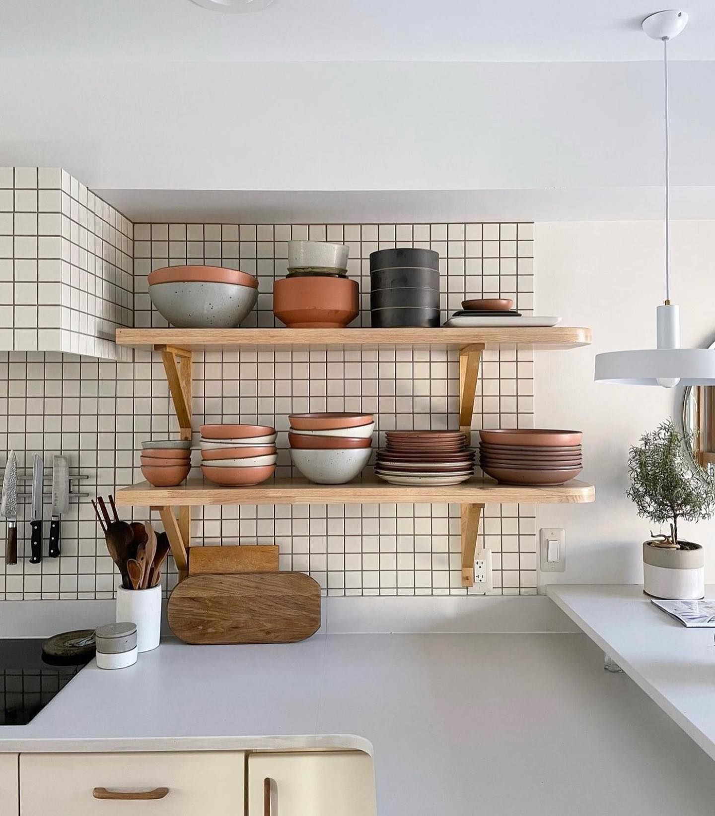 In a clean bright kitchen, there are open wooden shelves filled with ceramic plates and bowls against a grid tile