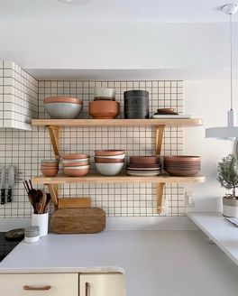In a clean bright kitchen, there are open wooden shelves filled with ceramic plates and bowls against a grid tile