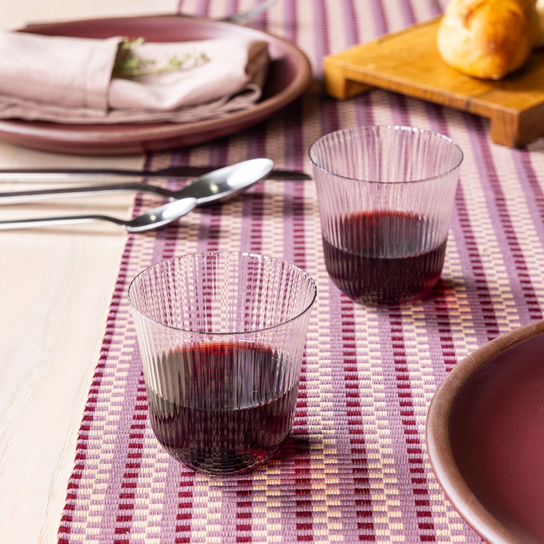 2 ribbed glasses that curve out on top filled with red wine and sitting on a striped table runner.