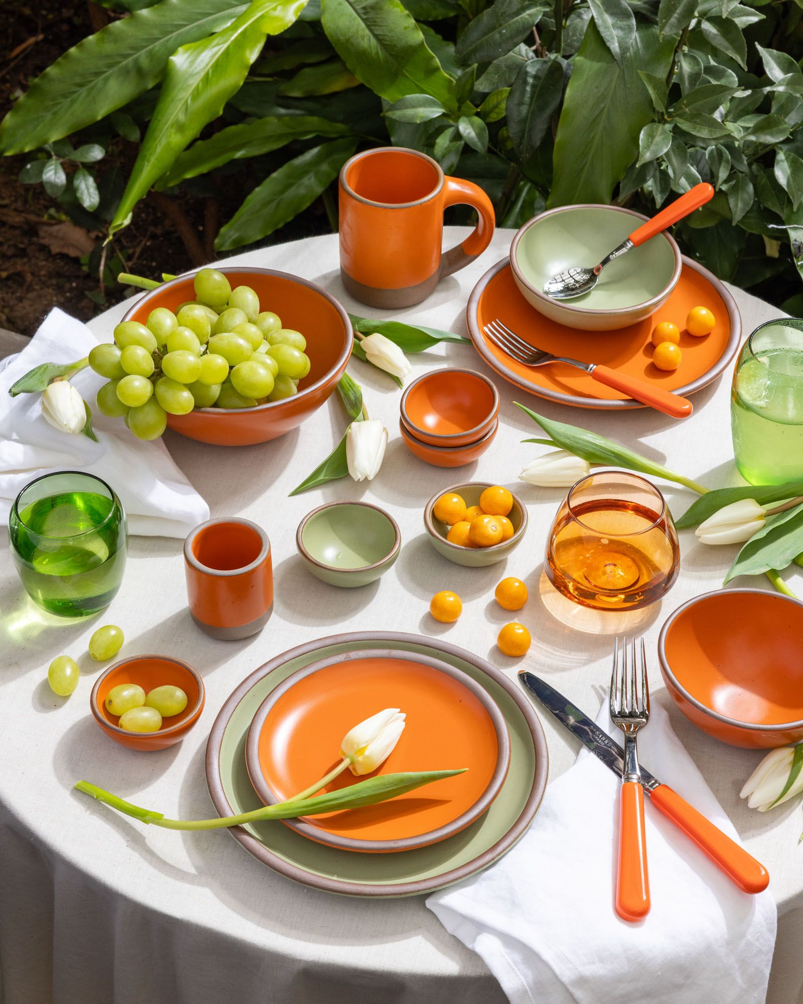 A table is covered in bold orange and calming sage green dinnerware, glasses, flatware and flowers in the same color palette.