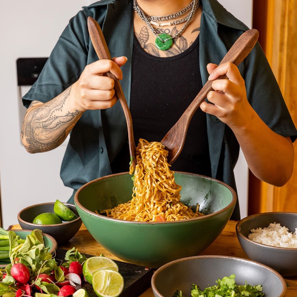 In a kitchen setting, a person holds two long walnut wood serving utensils that are grabbing a noodle dish from a large ceramic bowl. Around the bowl are other small bowls filled with chopped ingredients.