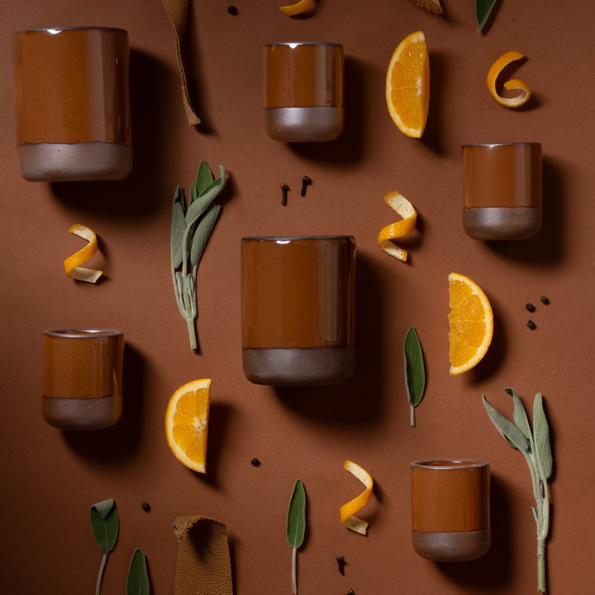 Glossy caramel brown ceramic tumblers arranged with orange slices, peel, sage leaves, and spices on a brown background.