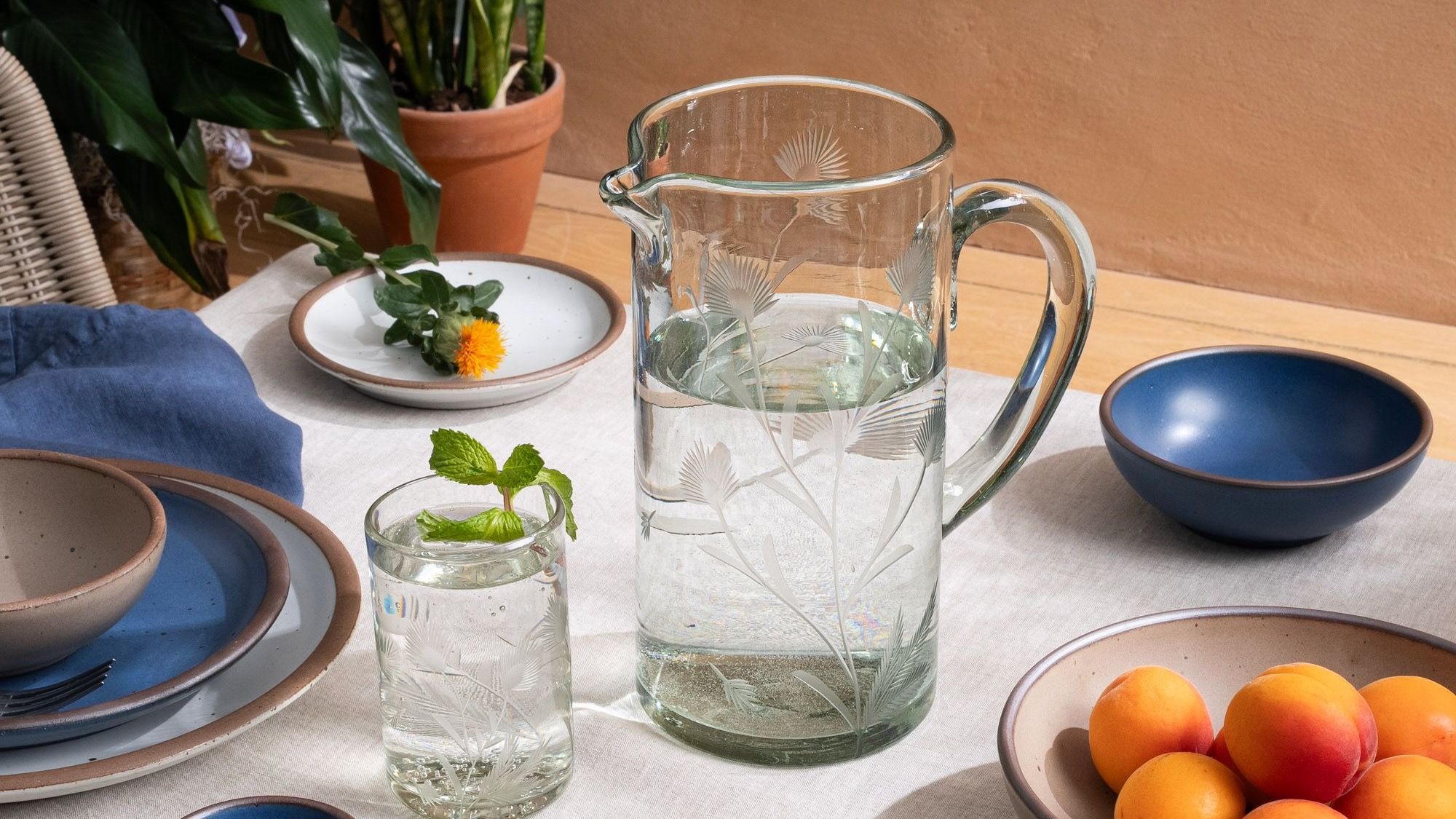 An etched thistle tumbler glass and pitcher filled with water, on a table with ceramic dinnerware and fruit, with plants in the background.