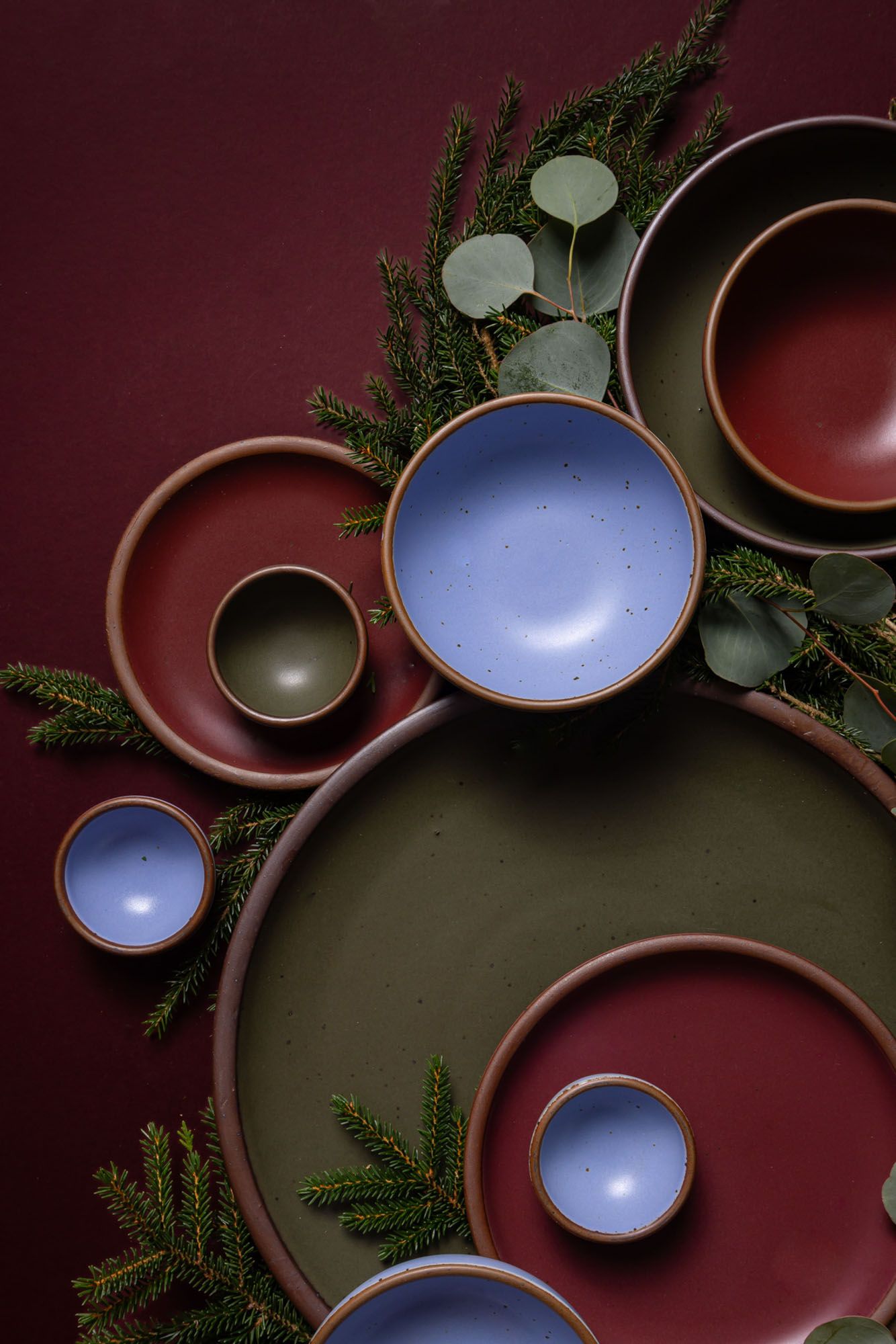 An artful overhead view of ceramic bowls and plates in plum, periwinkle, and forest green with eucalyptus and pine against a plum background.