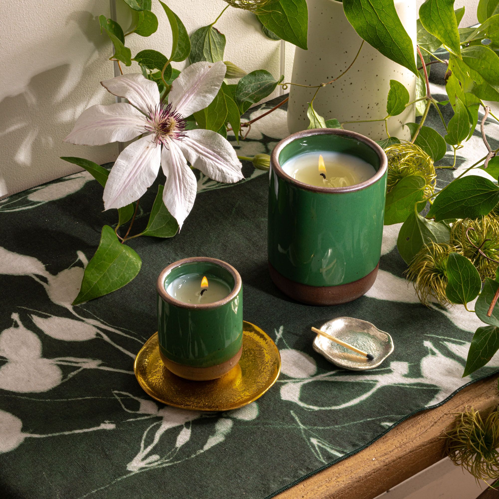 Two glossy emerald green ceramic candles on a floral cloth with a large white bloom, greenery, and a small dish holding a match.