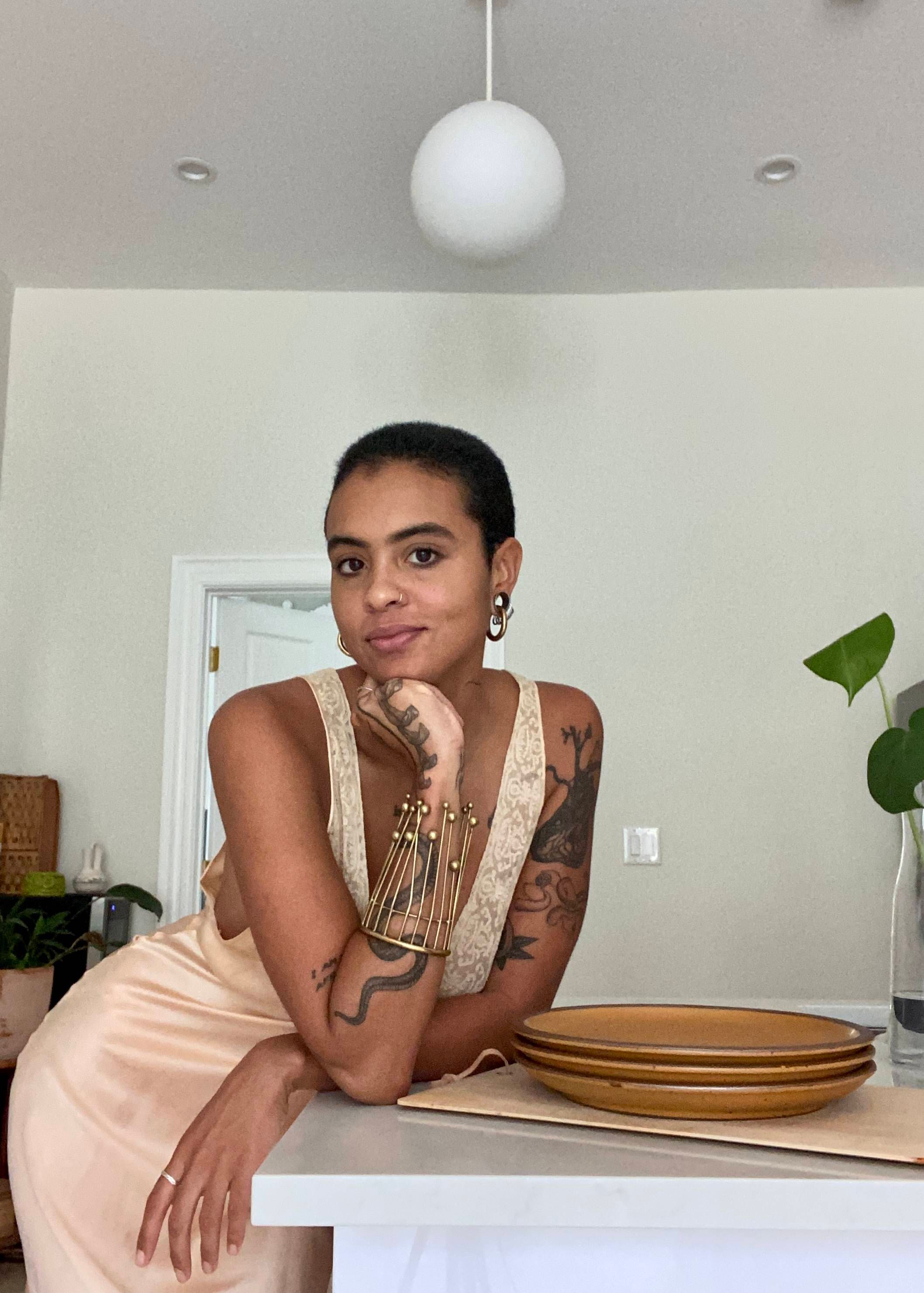 A person wearing a satin and lace dress leans on a counter with a stack of golden ceramic plates.