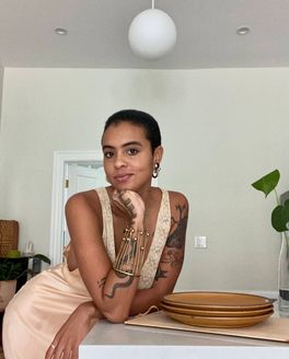 A person wearing a satin and lace dress leans on a counter with a stack of golden ceramic plates.