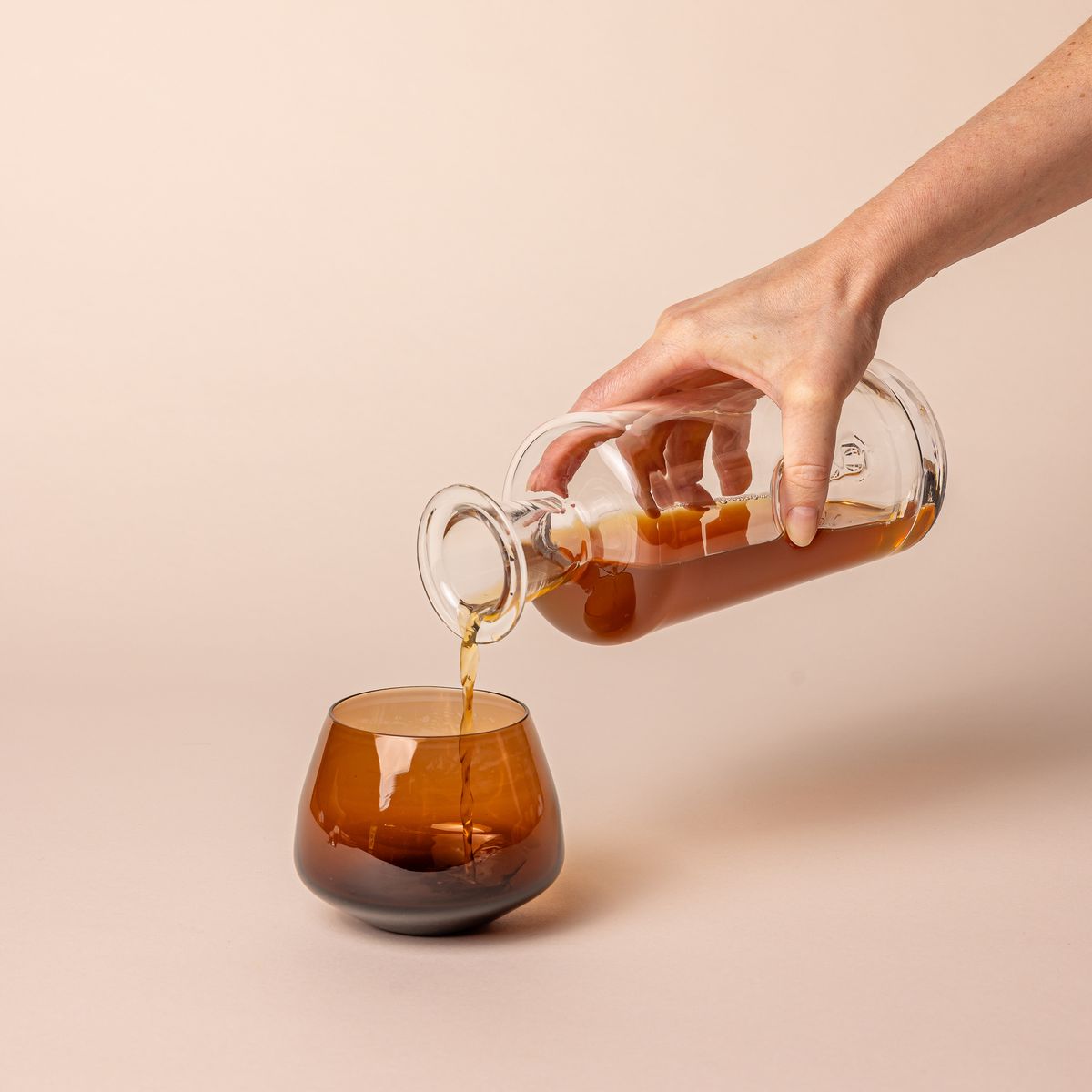 A hand holding a clear glass decanter of whiskey and pouring it into a brown glass whiskey snifter with a rounded base