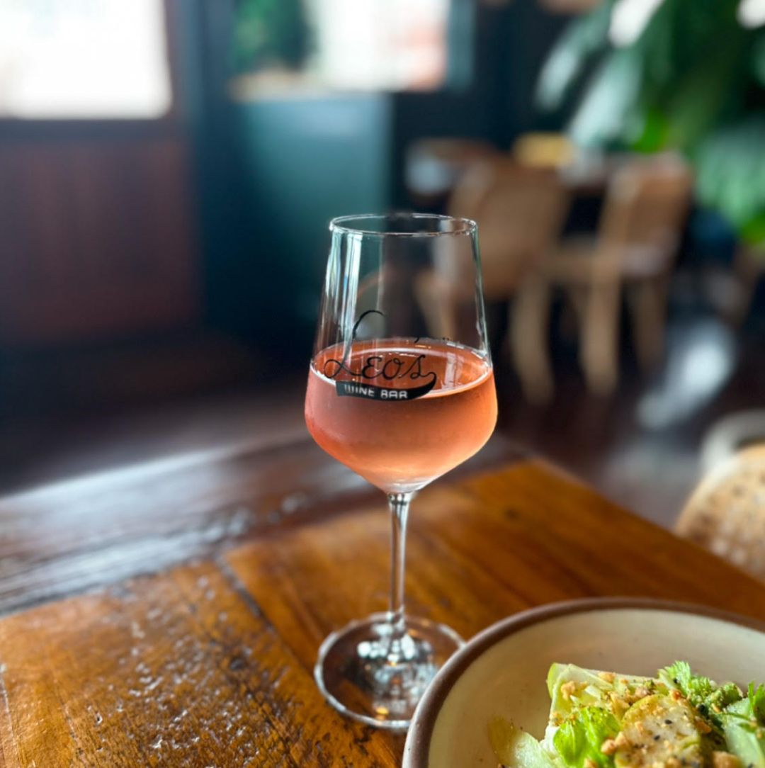 A glass of rosé wine on a wooden table at Leo’s Wine Bar, with a plate of salad partially visible in the foreground.