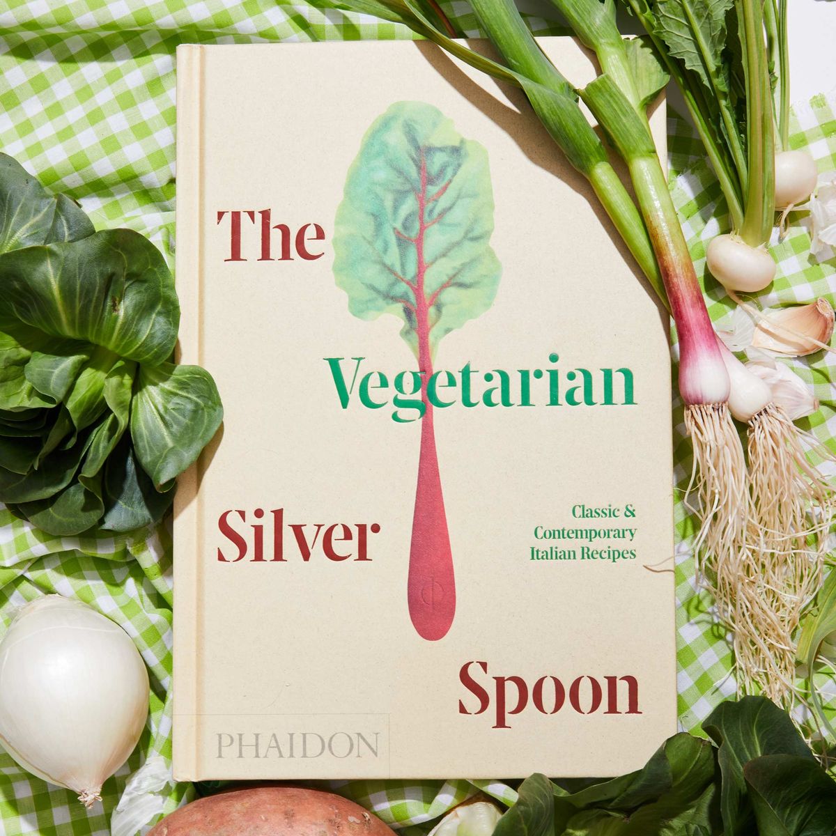 Vegetables are on either side and above and below A hand holds the top of the book The Vegetarian Silver Spoon, which has an illustration of a piece of Swiss chard