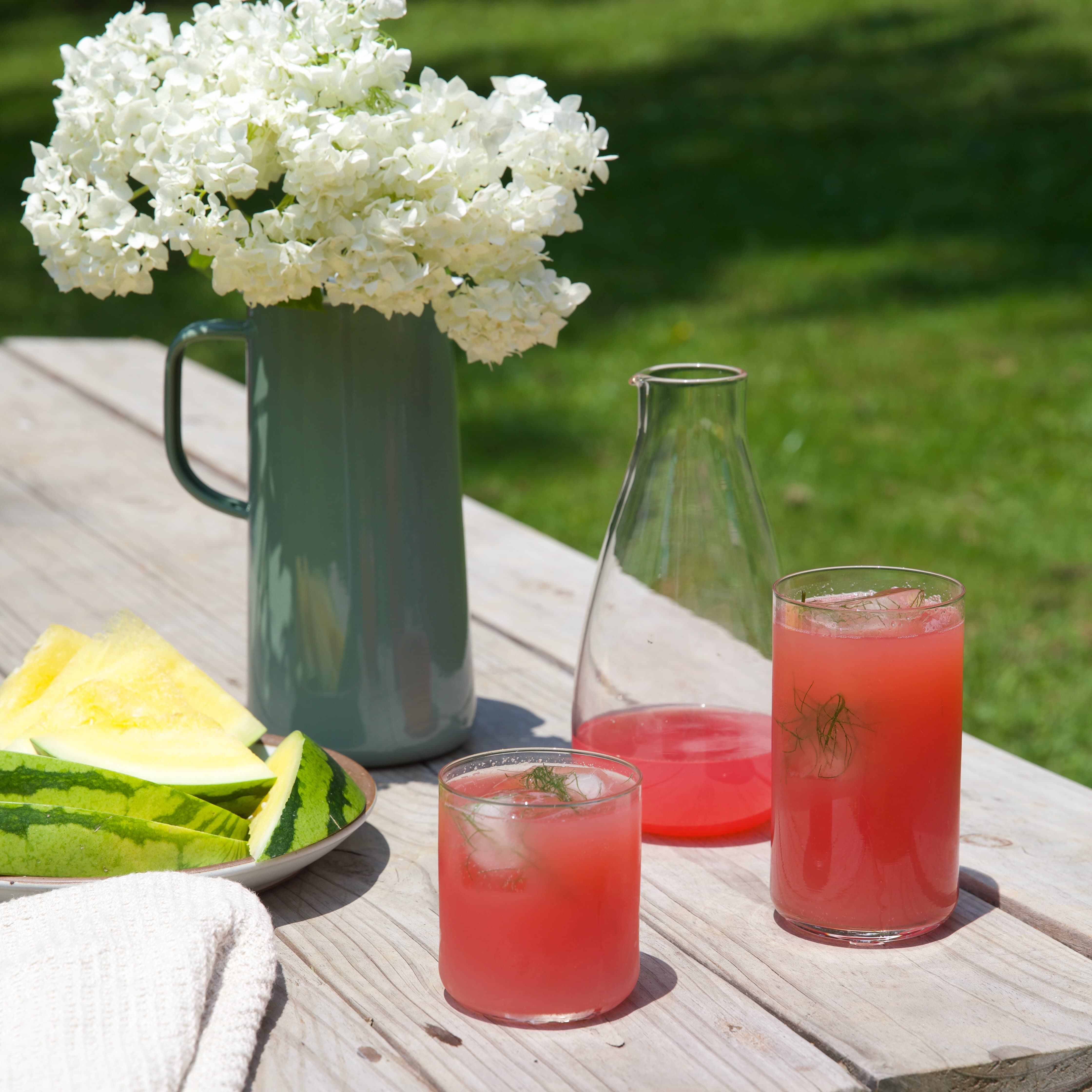 On a picnic table is 2 glasses and a carafe filled with a pink drink with ice. There is also a plate of sliced watermelon, and a pitcher with flowers.