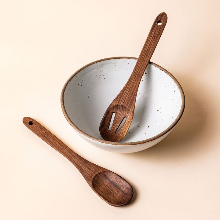 1 large slotted wooden spoon sits inside a large white ceramic mixing bowl, and a large everyday mixing spoon sits styled beside it.