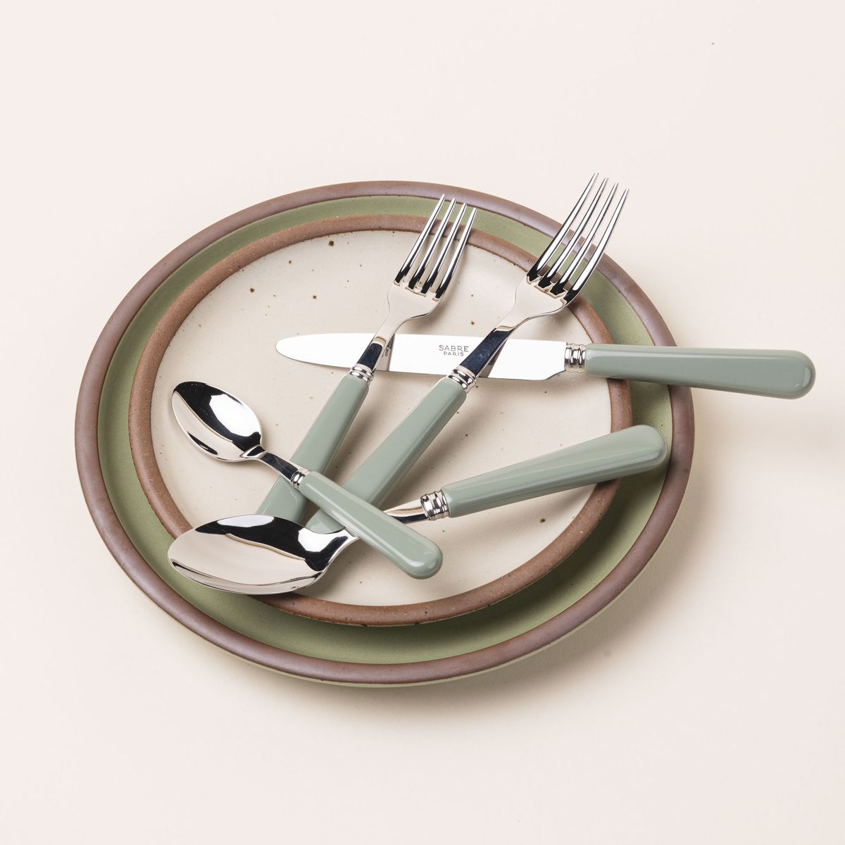 A five piece flatware set with shiny utensils and matte sage green handles sits on two ceramic plates in off-white and calming sage green