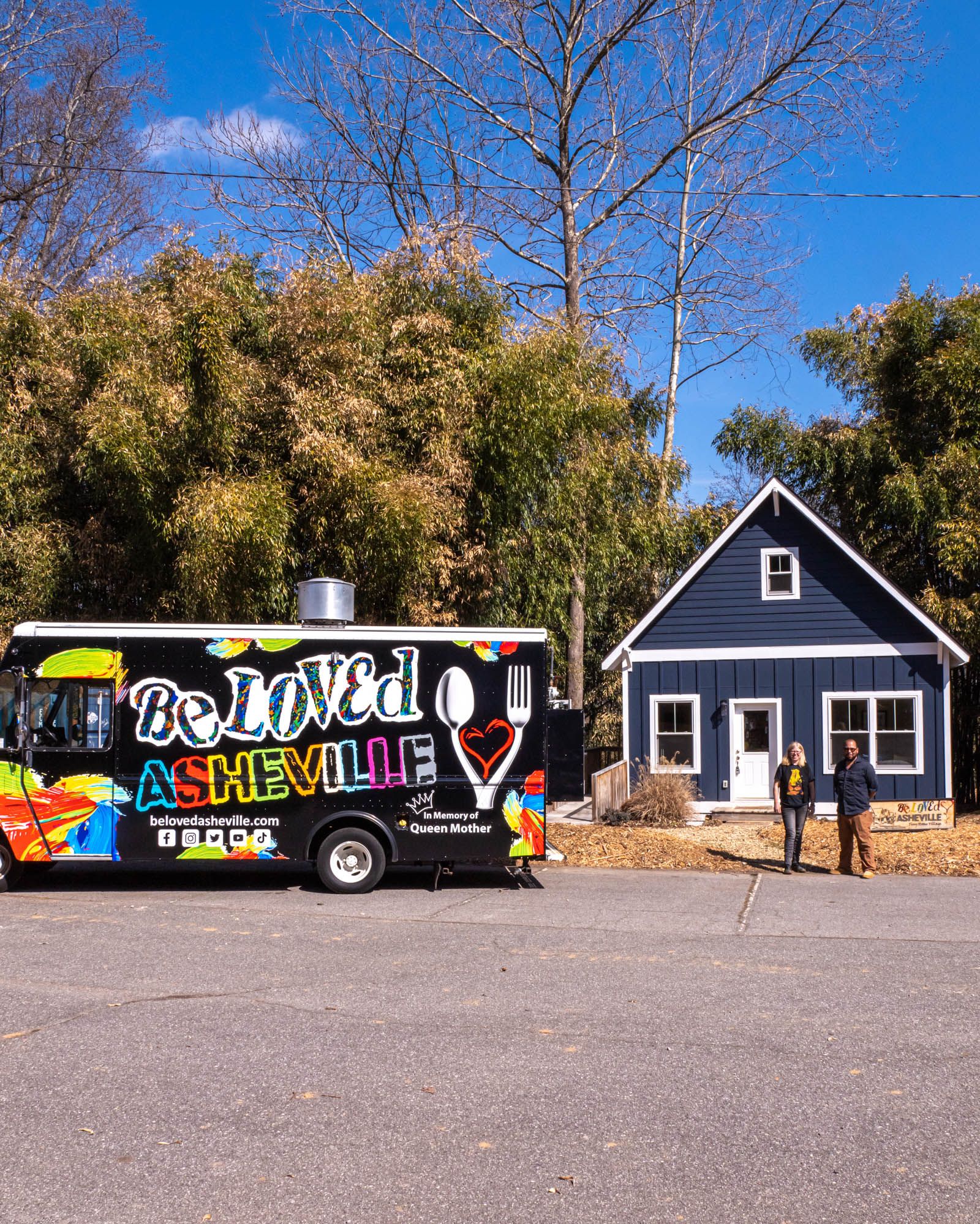 A large truck that reads "BeLoved Asheville" sits outside a navy home with 2 people standing outside.