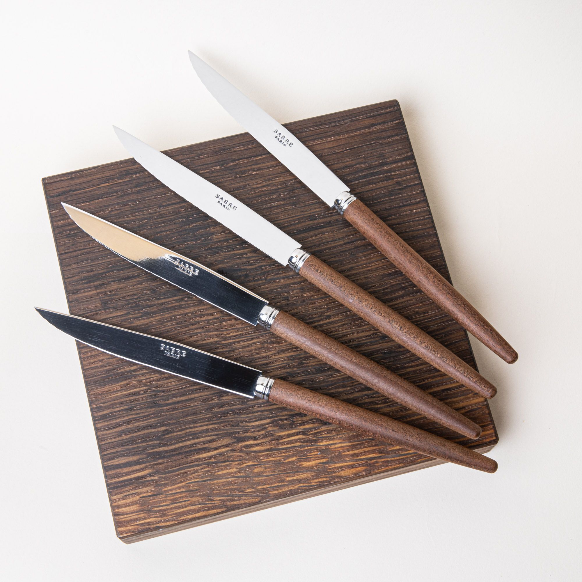 Four steak knives with steel blades and simple wood handles, fanned over a dark wood cutting board