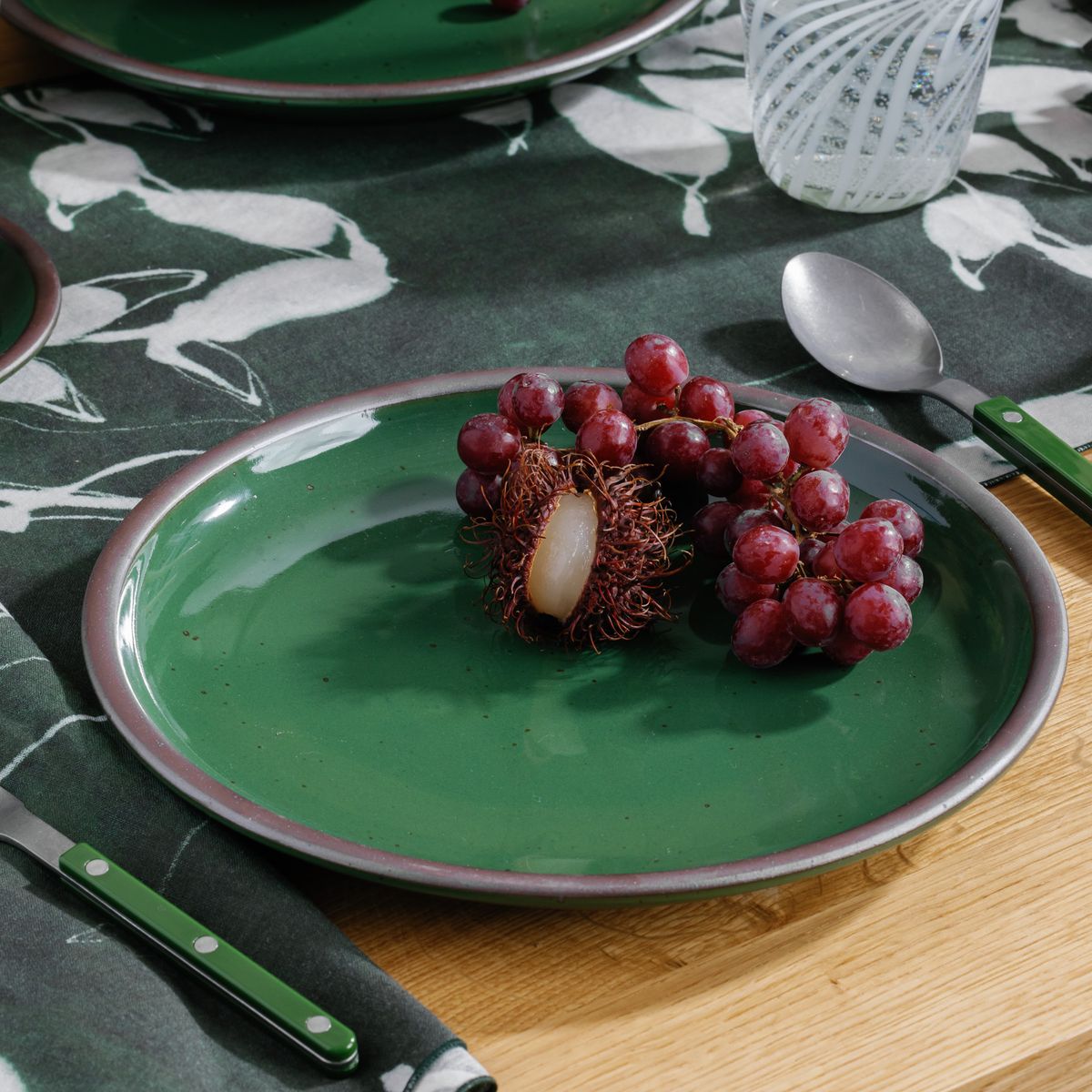 Glossy emerald green ceramic plate with clusters of red grapes on a table with a leafy patterned cloth.