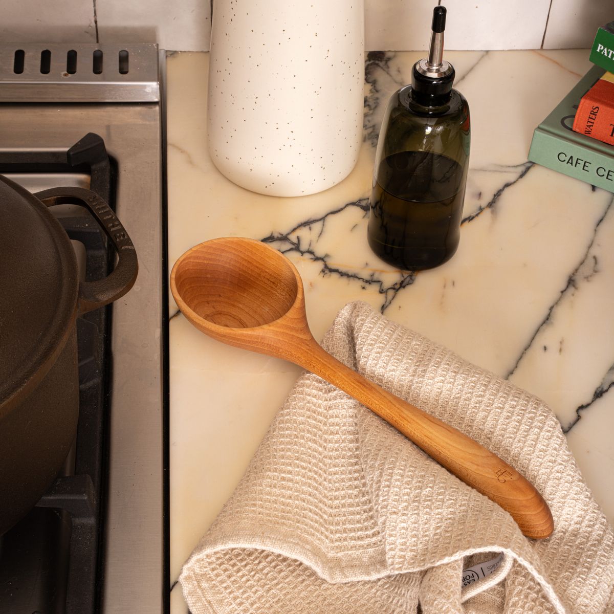 A wooden ladle with a large scoop next to a stovetop with a waffle weave kitchen towel and cruet.