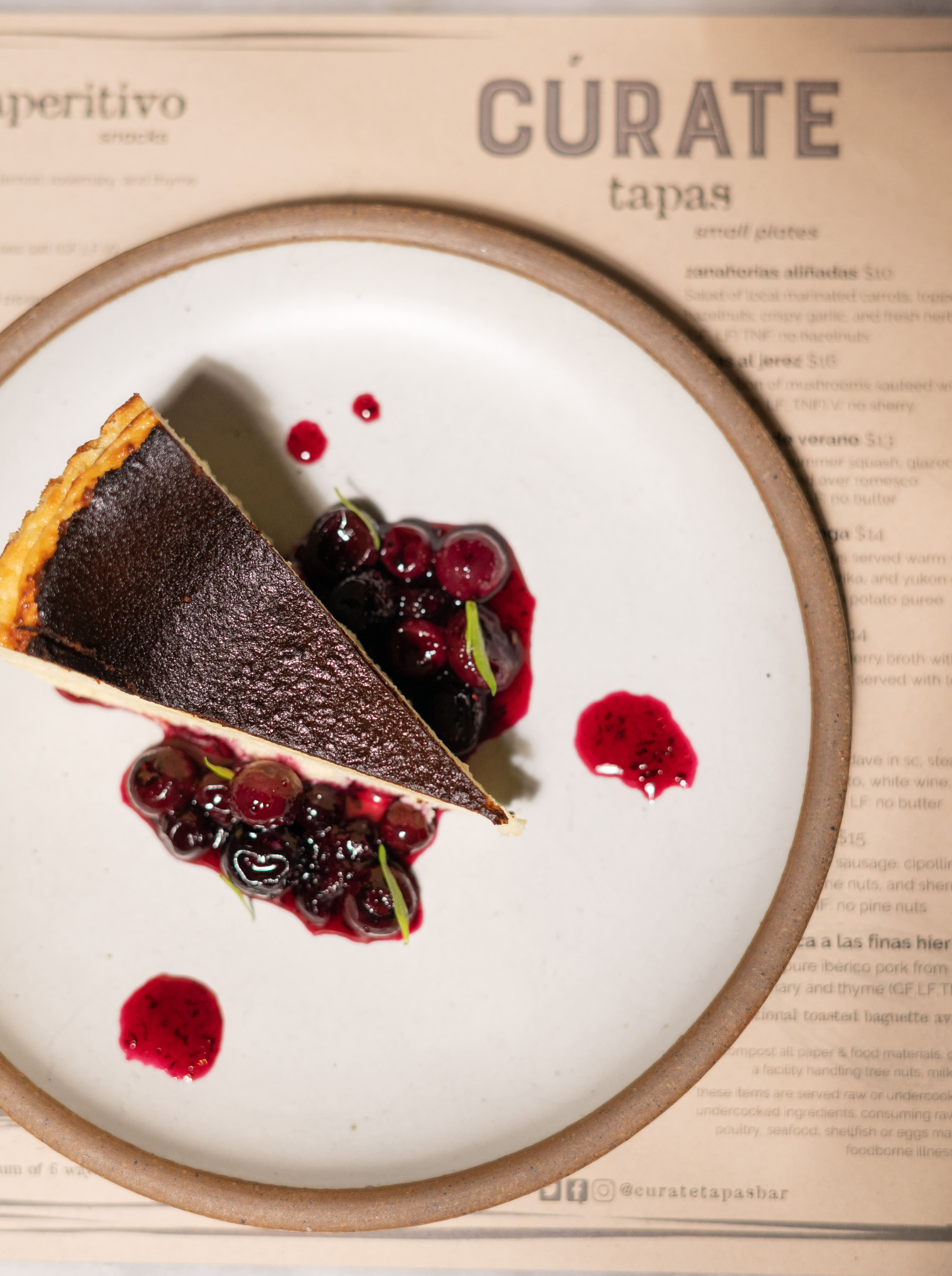 Slice of cheesecake plated on East Fork Eggshell plate with berry mixture. Plate is sitting on top of a Curate menu