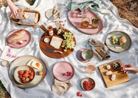 An outdoor picnic filled with ceramic dinnerware in dusty mauve pink and grey-green colors, bread, berries and cheese, a charcuterie spread, glassware and more.