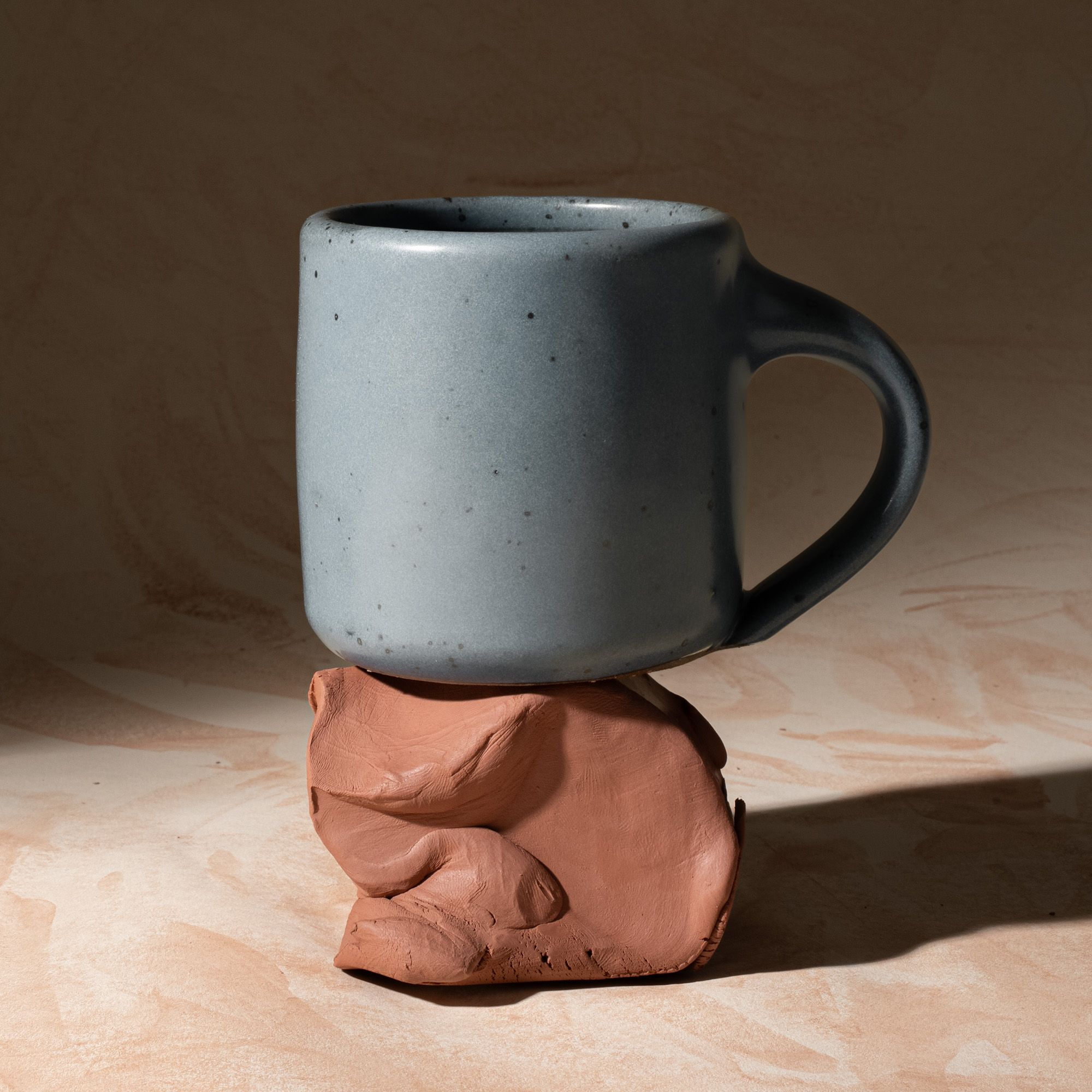 A medium sized ceramic mug with handle in a light blue-grey color featuring iron speckles, sitting on a block of raw clay.