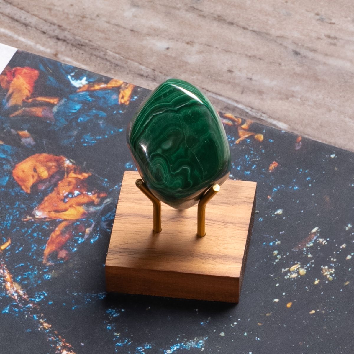 Polished green malachite stone on brass stand with wooden base, displayed against a colorful, abstract book cover.