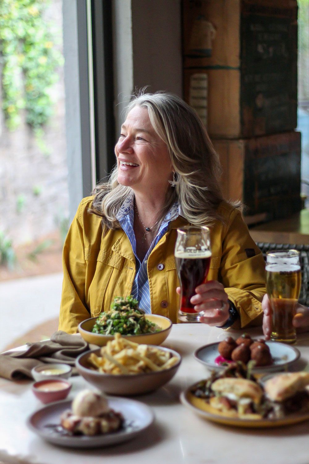 A person is smiling and sits at a table filled with various plates of foods including fries, salad, a sandwich, and dessert.