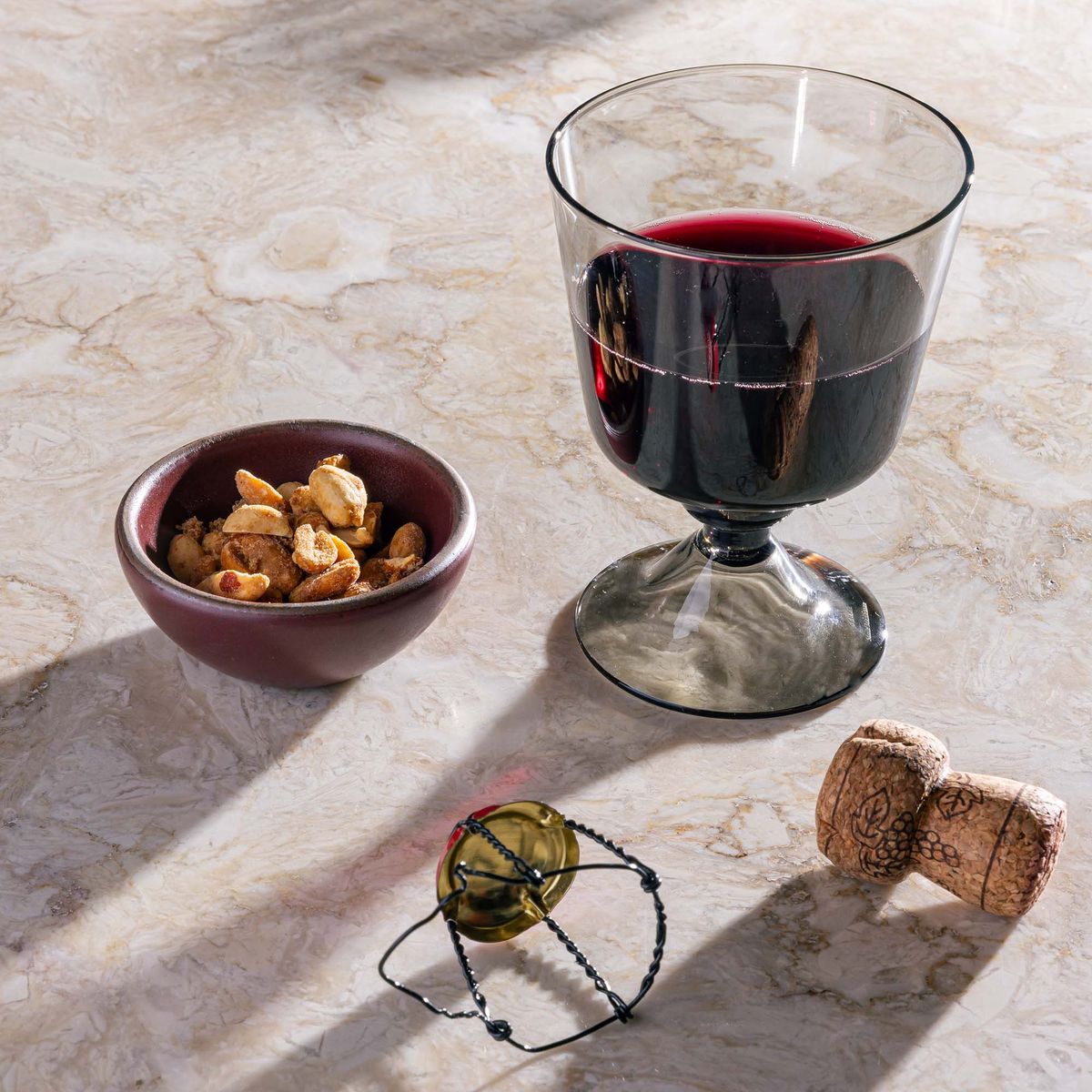 A short wine glass with an angular cup and grey color filled with red wine, with a tiny bowl of nuts, and a cork