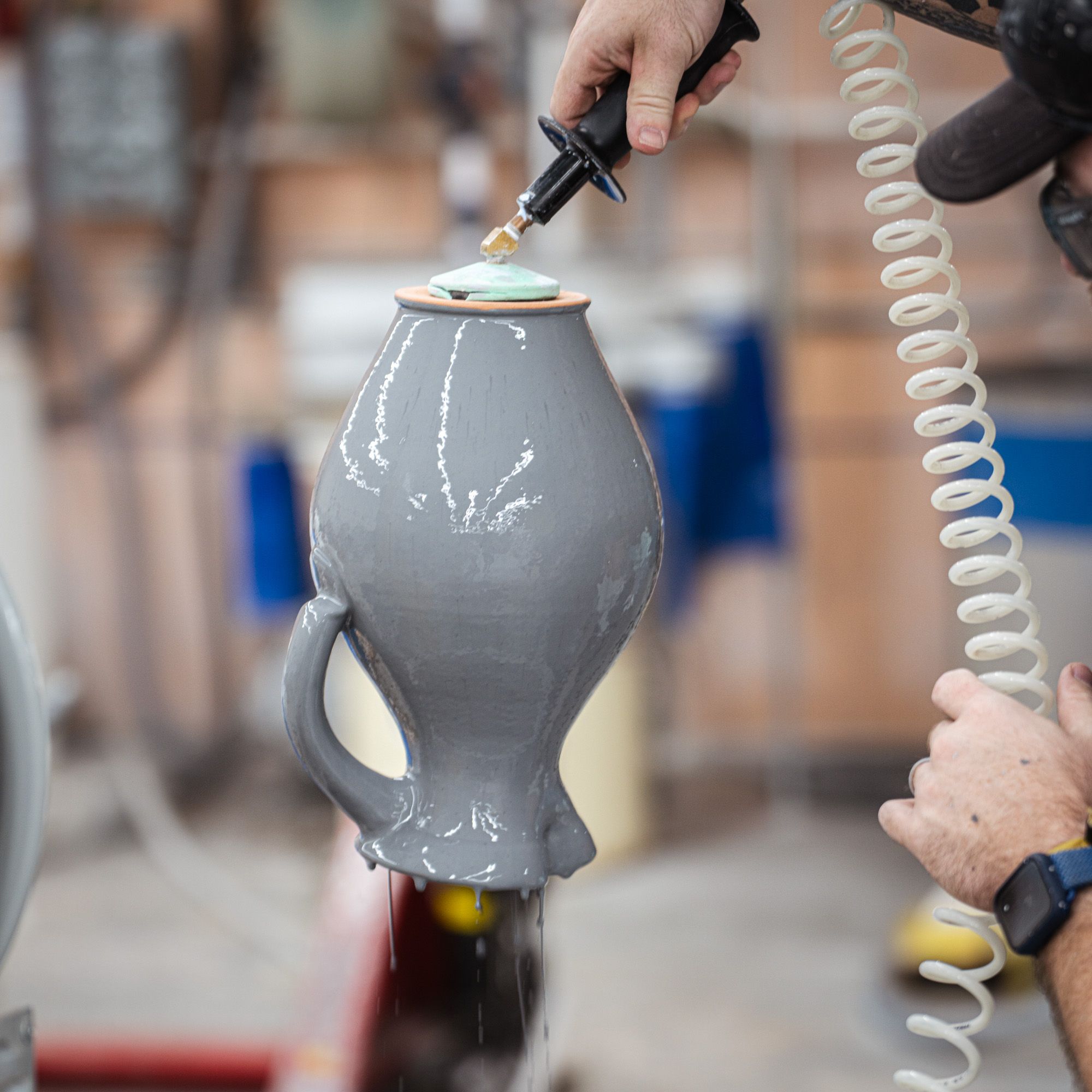 Hands holding a freshly glazed gray pitcher upside down with a tool with glaze dripping off.