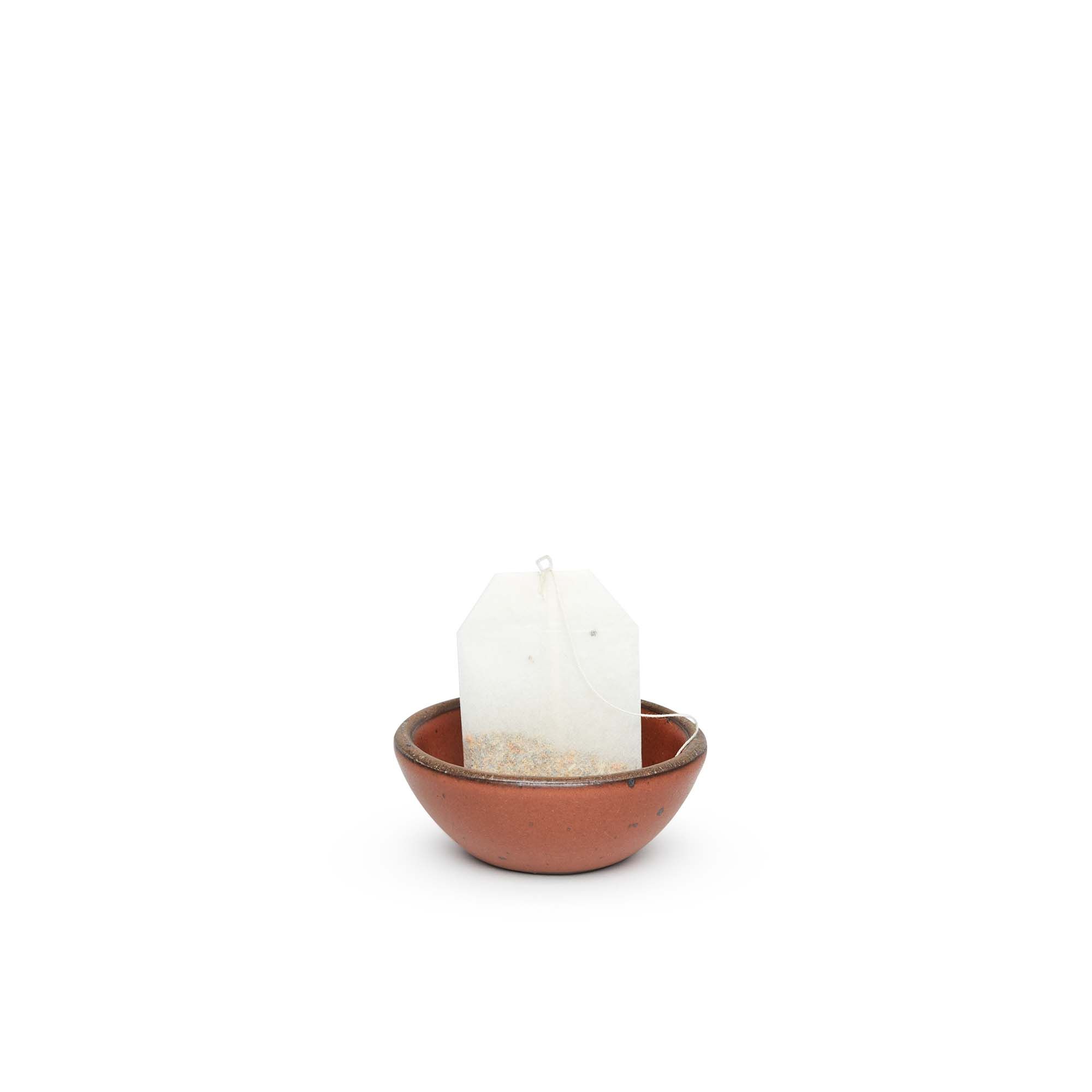A tiny rounded ceramic bowl in a cool burnt terracotta color featuring iron speckles and an unglazed rim, holding a tea bag