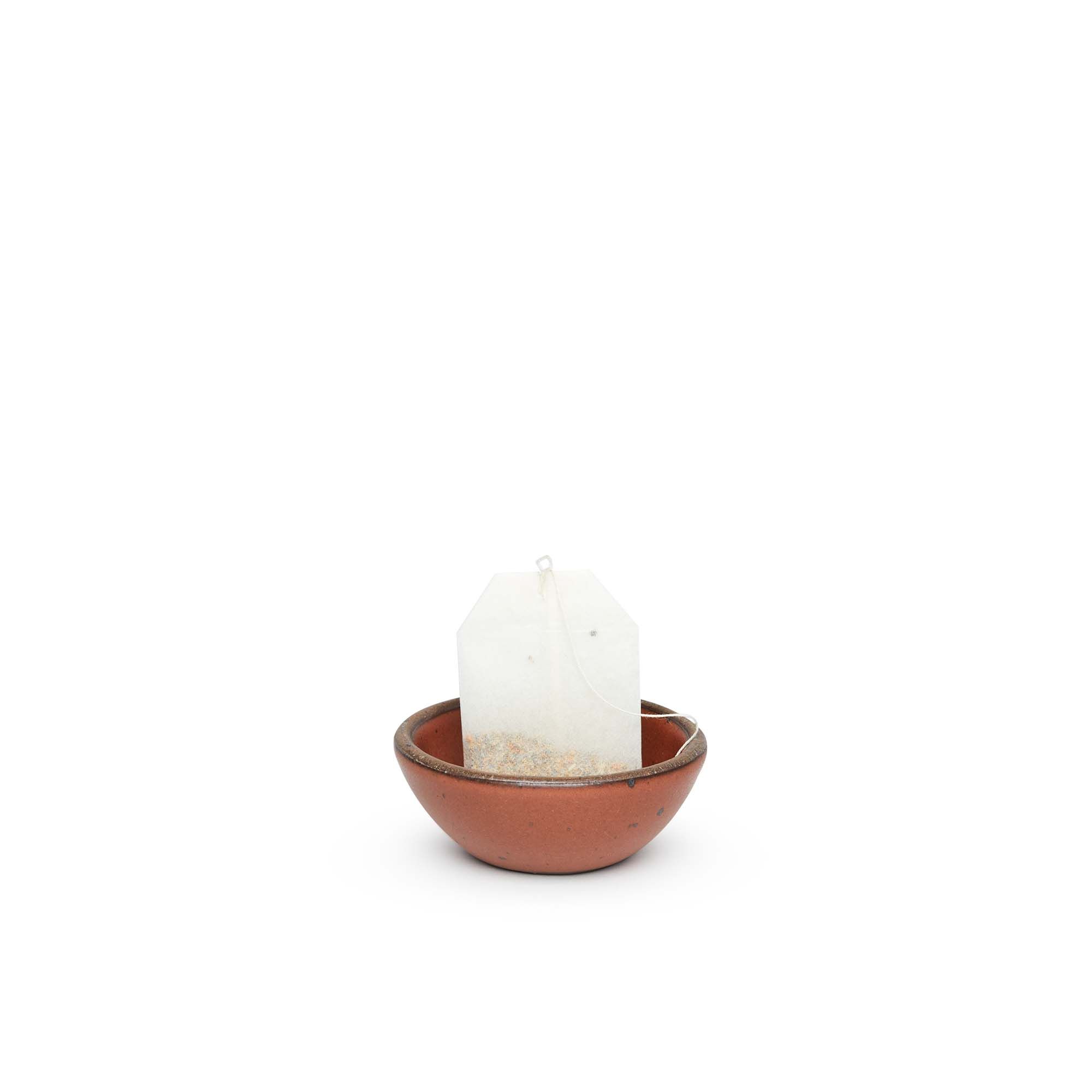 A tiny rounded ceramic bowl in a cool burnt terracotta color featuring iron speckles and an unglazed rim, with a teabag