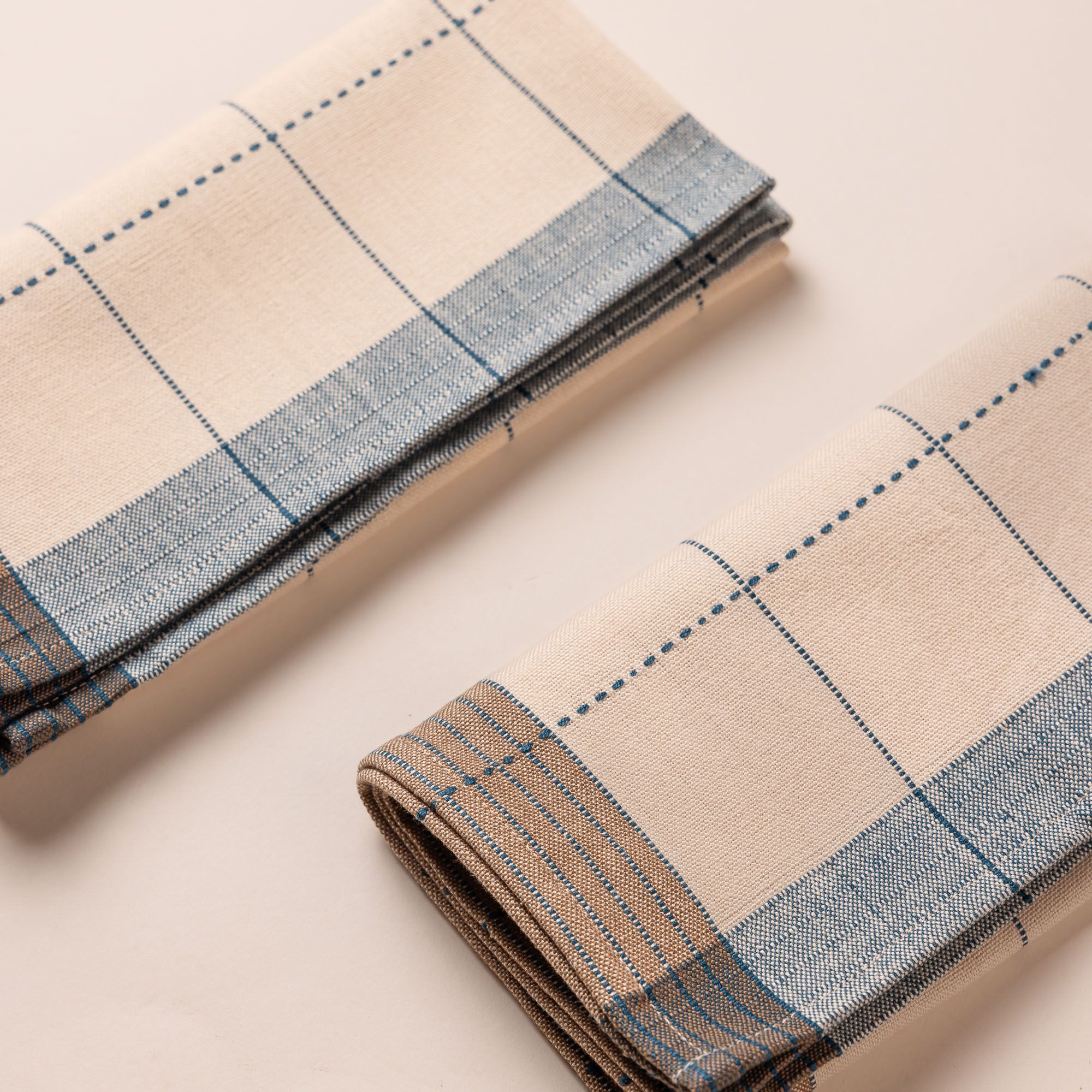 Two natural folded napkins side by side that are designed with turquoise gridlines with light blue and tan striped edging.