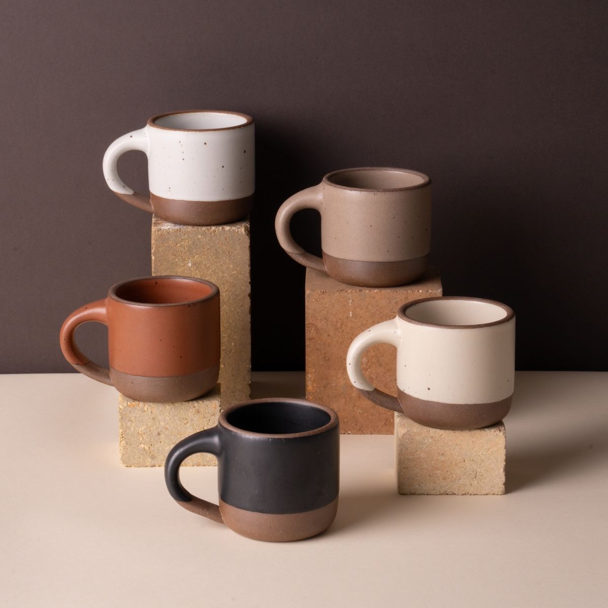 5 Small mugs are artfully arranged on different riser heights close together, in cool white, warm pale brown, terracotta, warm white, and graphite black colors.