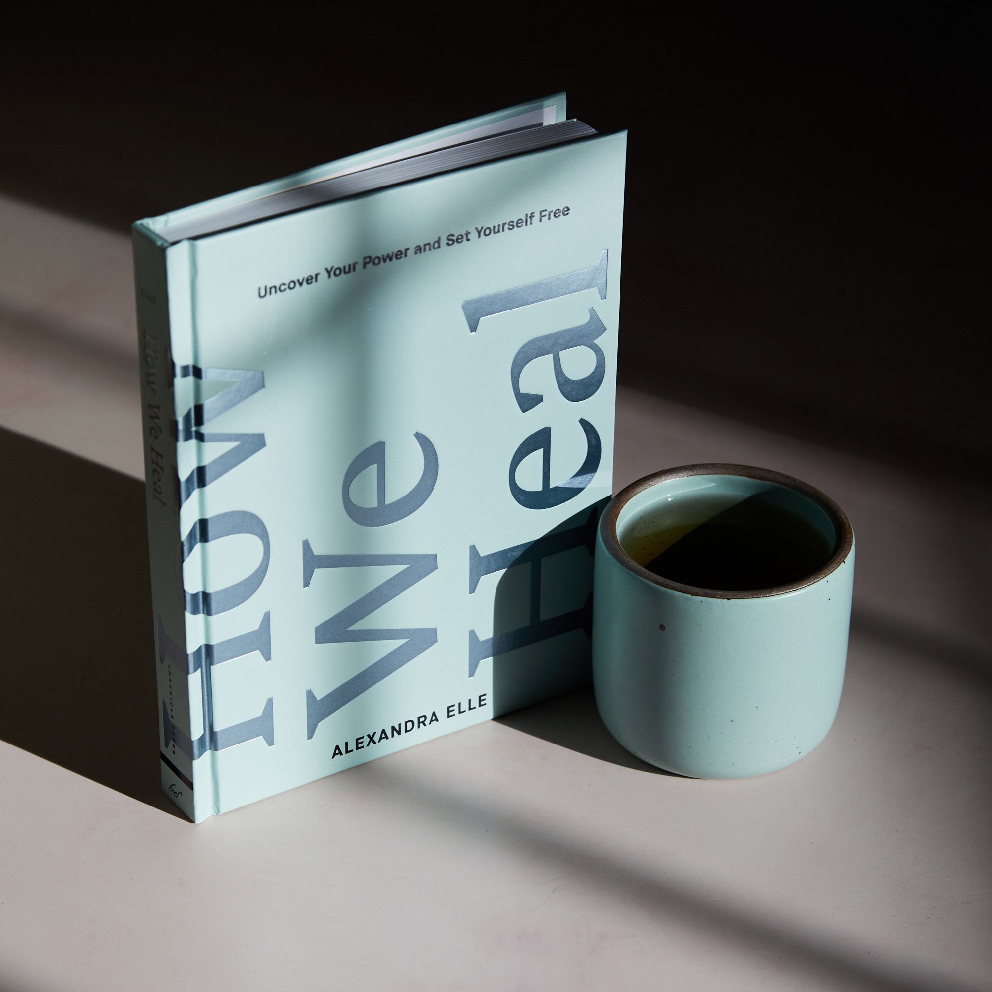 A book standing up right that reads 'How We Heal' and a ceramic cup in Malibu sitting beside it with shadows.